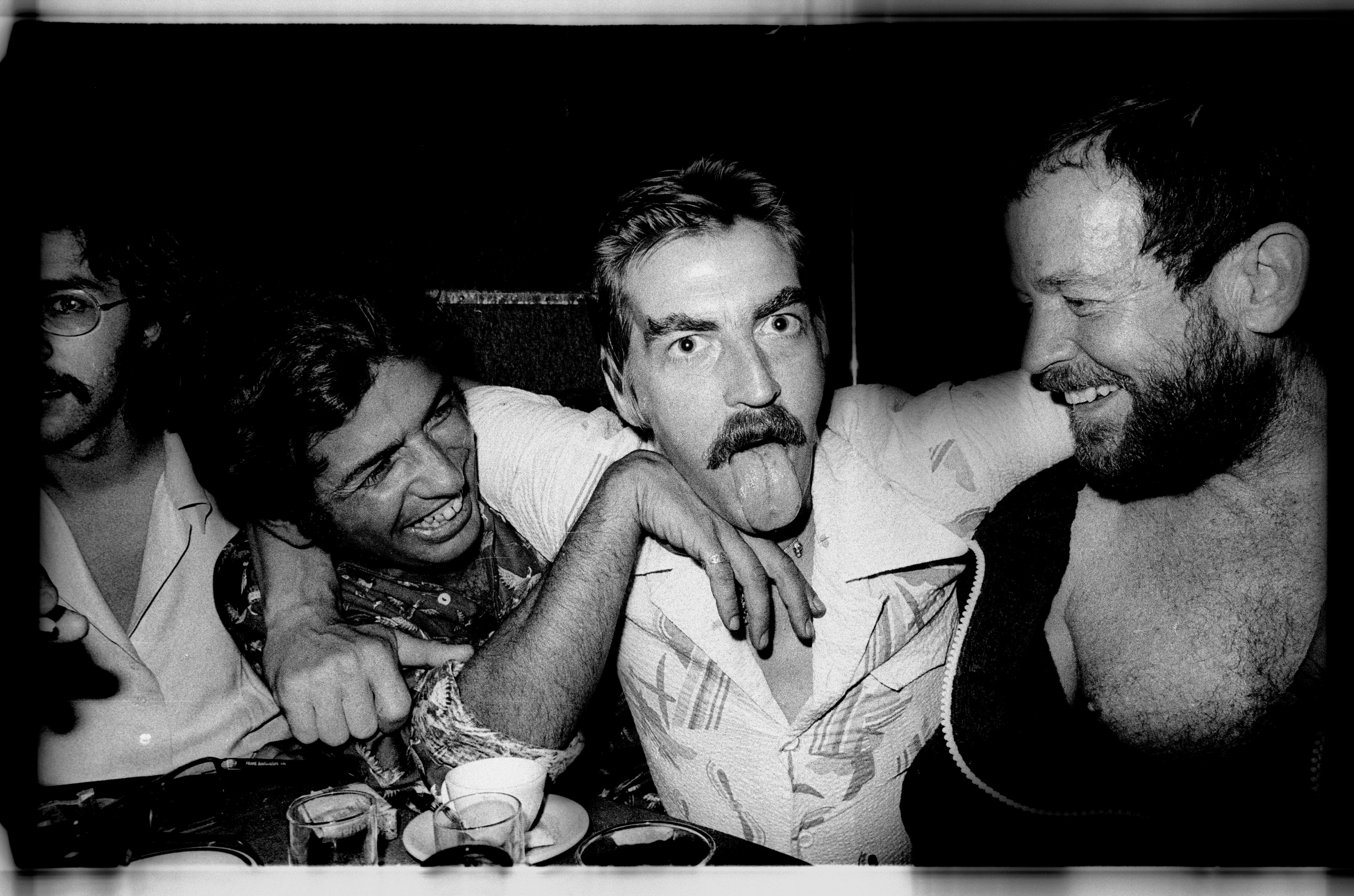 Four men, one with a moustache, laughing and embracing in a dimly lit room.