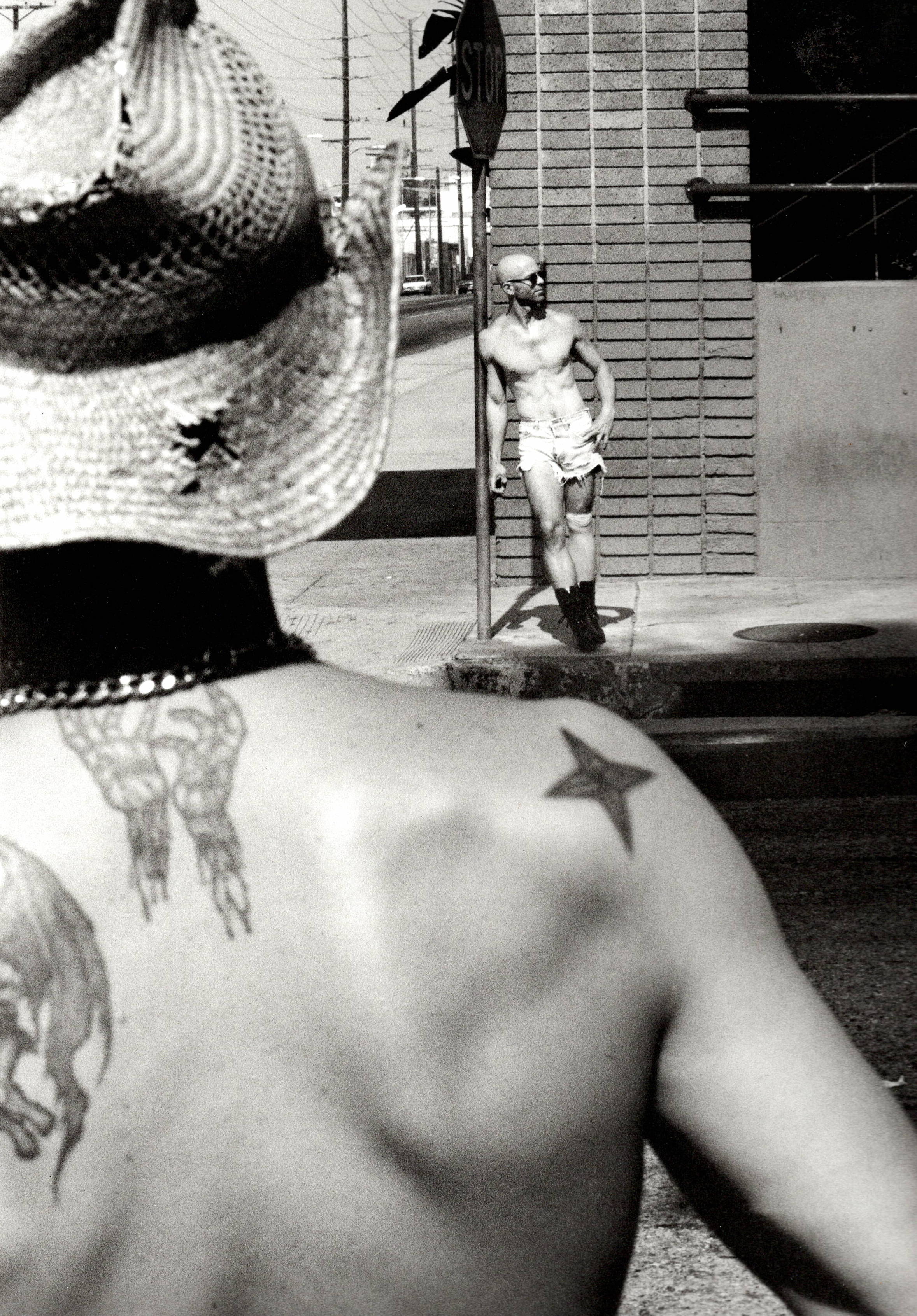 Black and white image showing tattooed person in straw hat in foreground, with figure standing by brick building and metal pole in background.