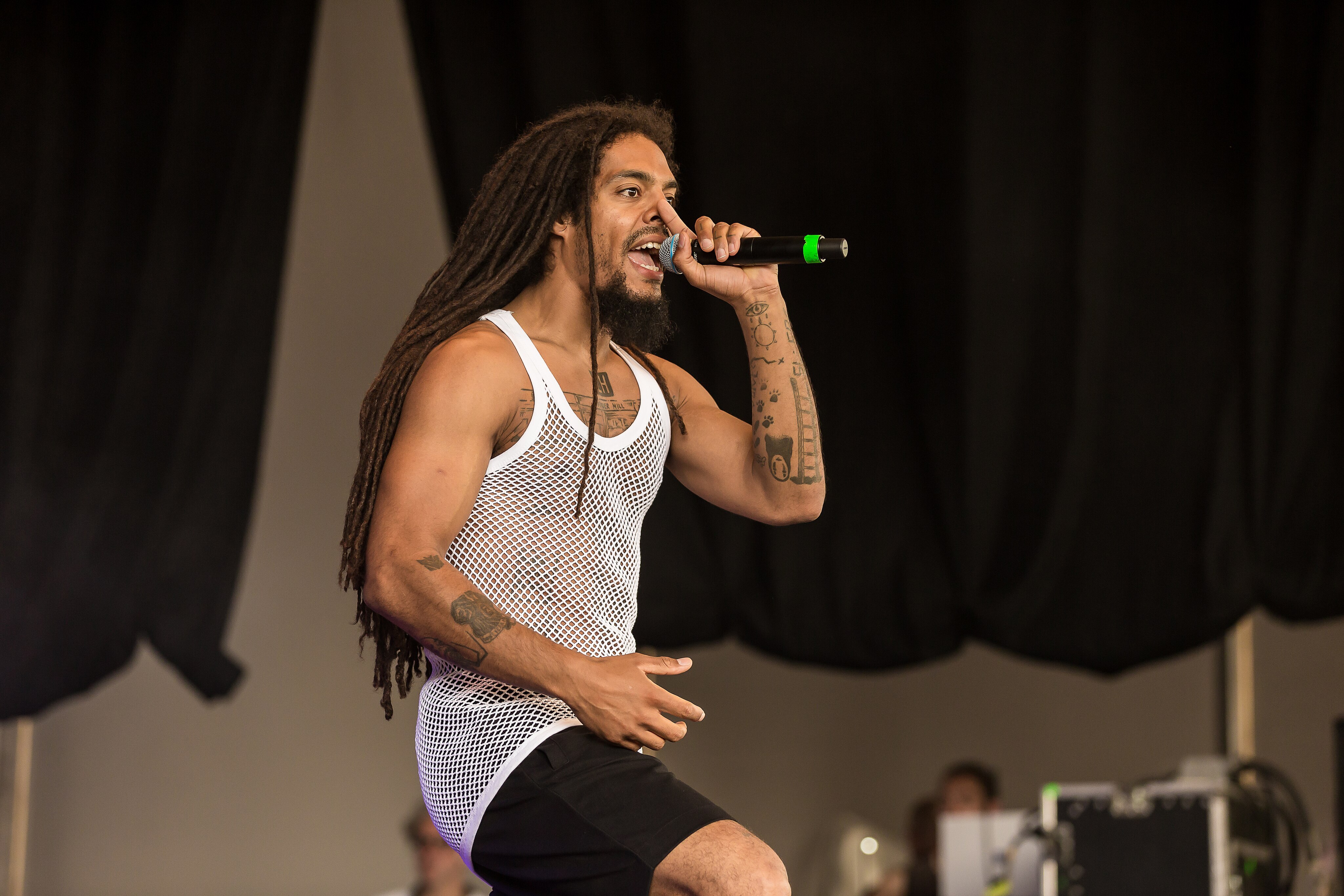 Performer with long dreadlocks singing into microphone, wearing white mesh vest and black shorts on stage.