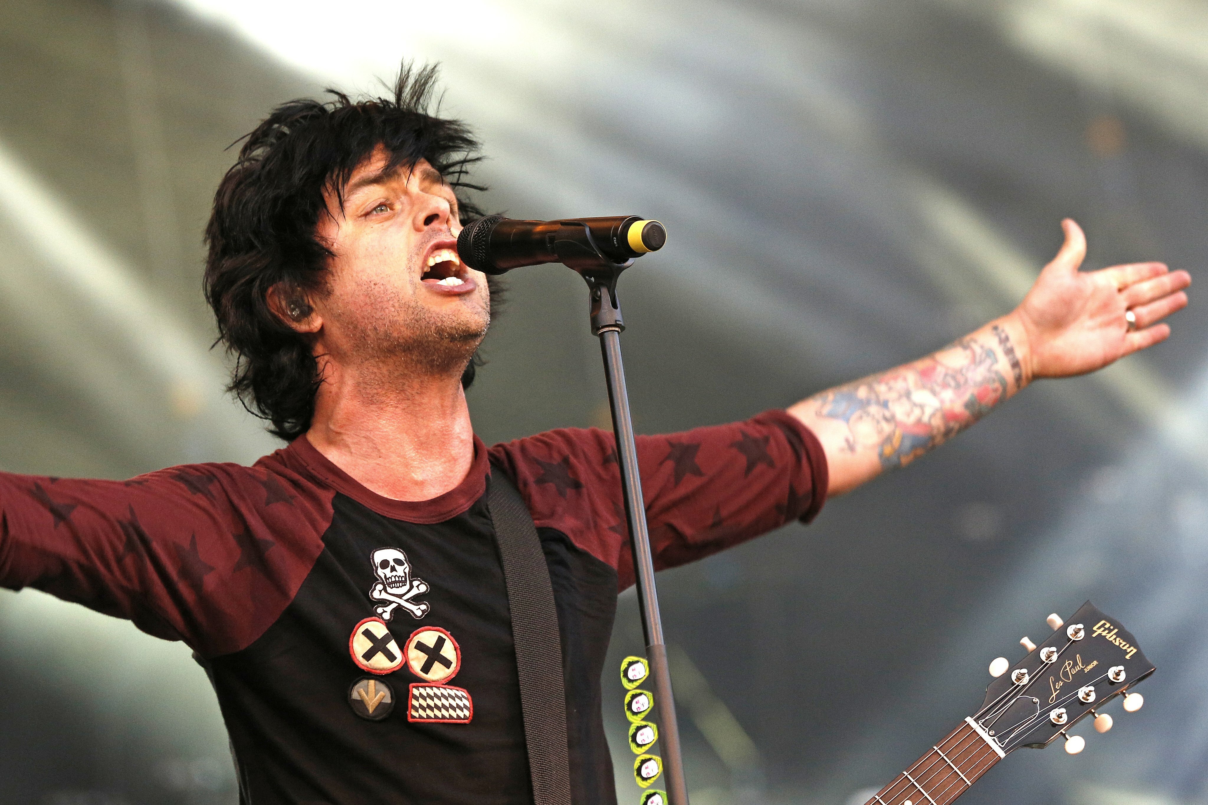 Man singing at microphone on stage, wearing red shirt and tattoo on arm, playing electric guitar.