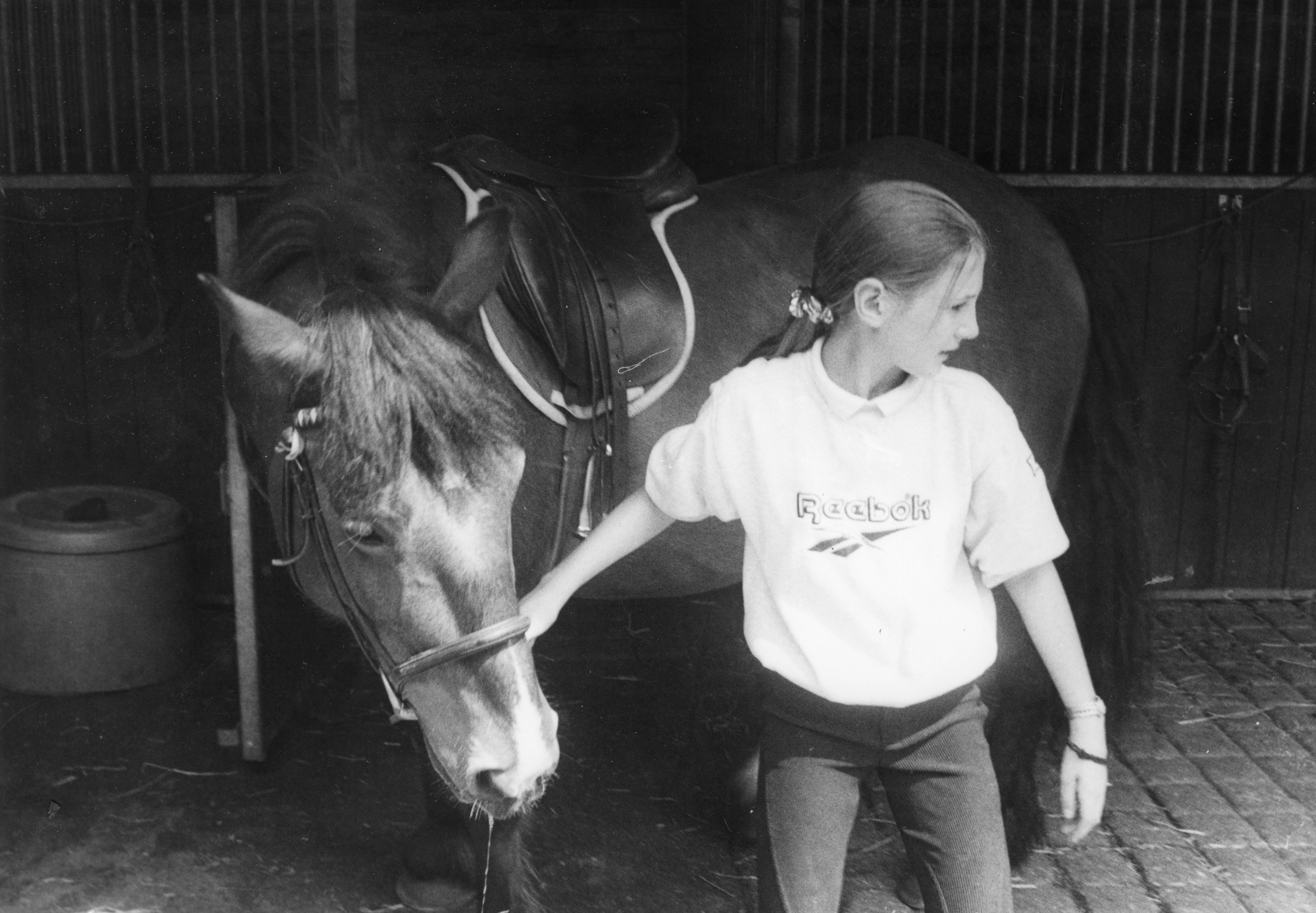 Two equine figures, a young human person standing beside a brown horse in a stable setting. The human is wearing a white t-shirt.