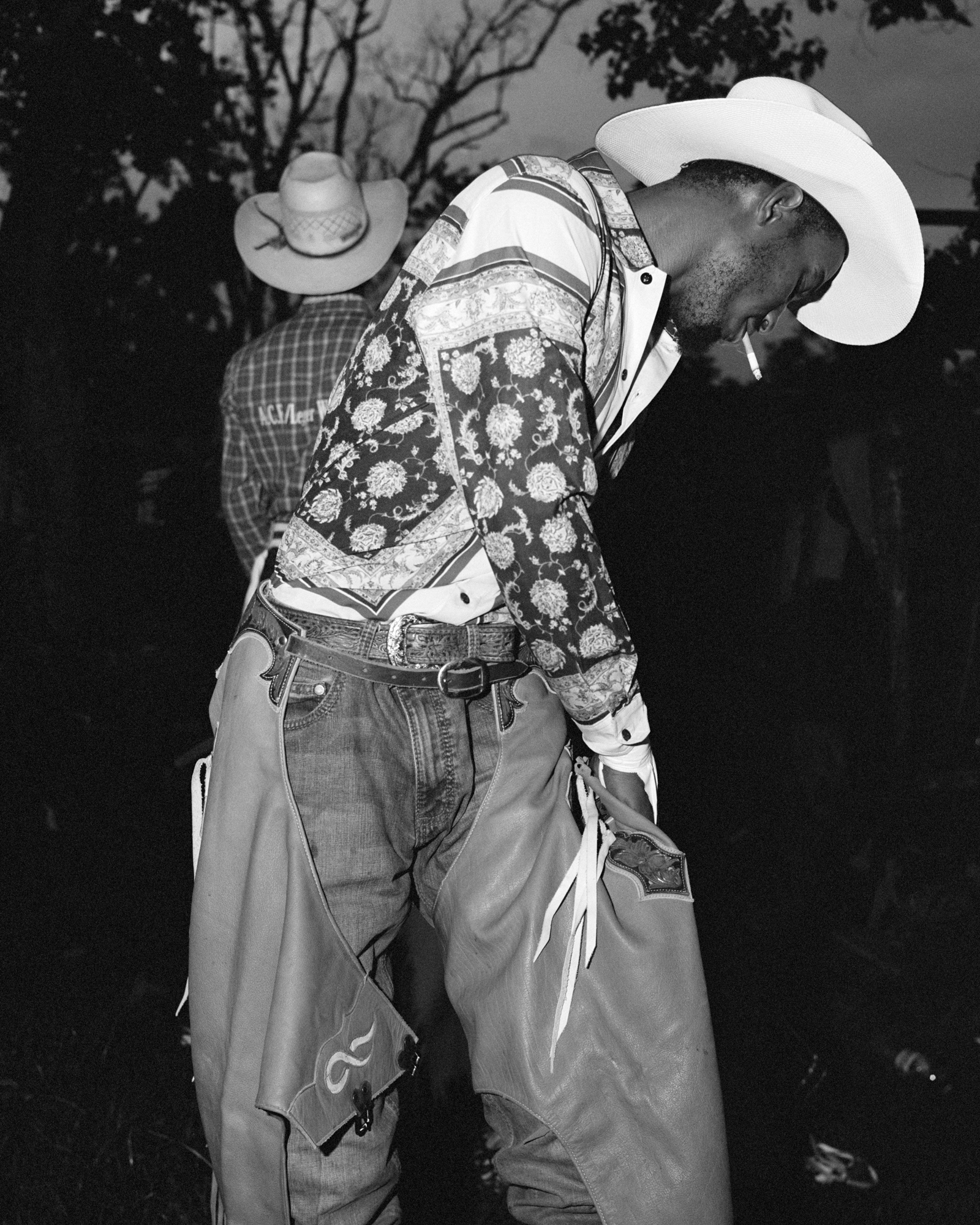 Two men wearing cowboy hats and Western-style clothing, one with a patterned shirt, standing outdoors in a natural setting.