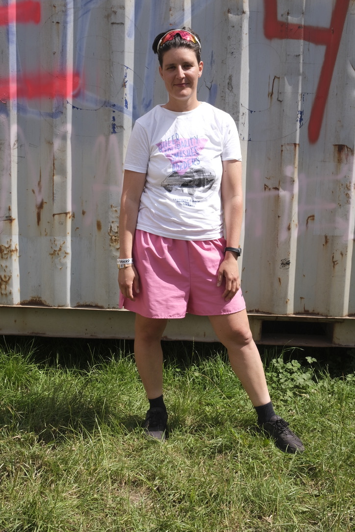 Woman in white t-shirt and pink shorts standing on grass in front of graffitied metal fence with red and blue markings.
