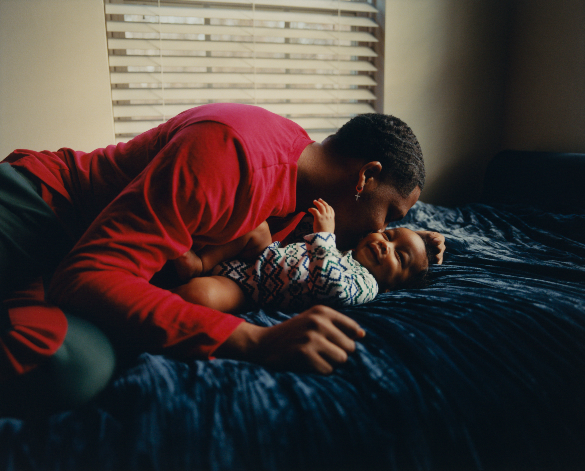 Father in red shirt leaning over infant on dark blue bedding, with white venetian blinds in background.