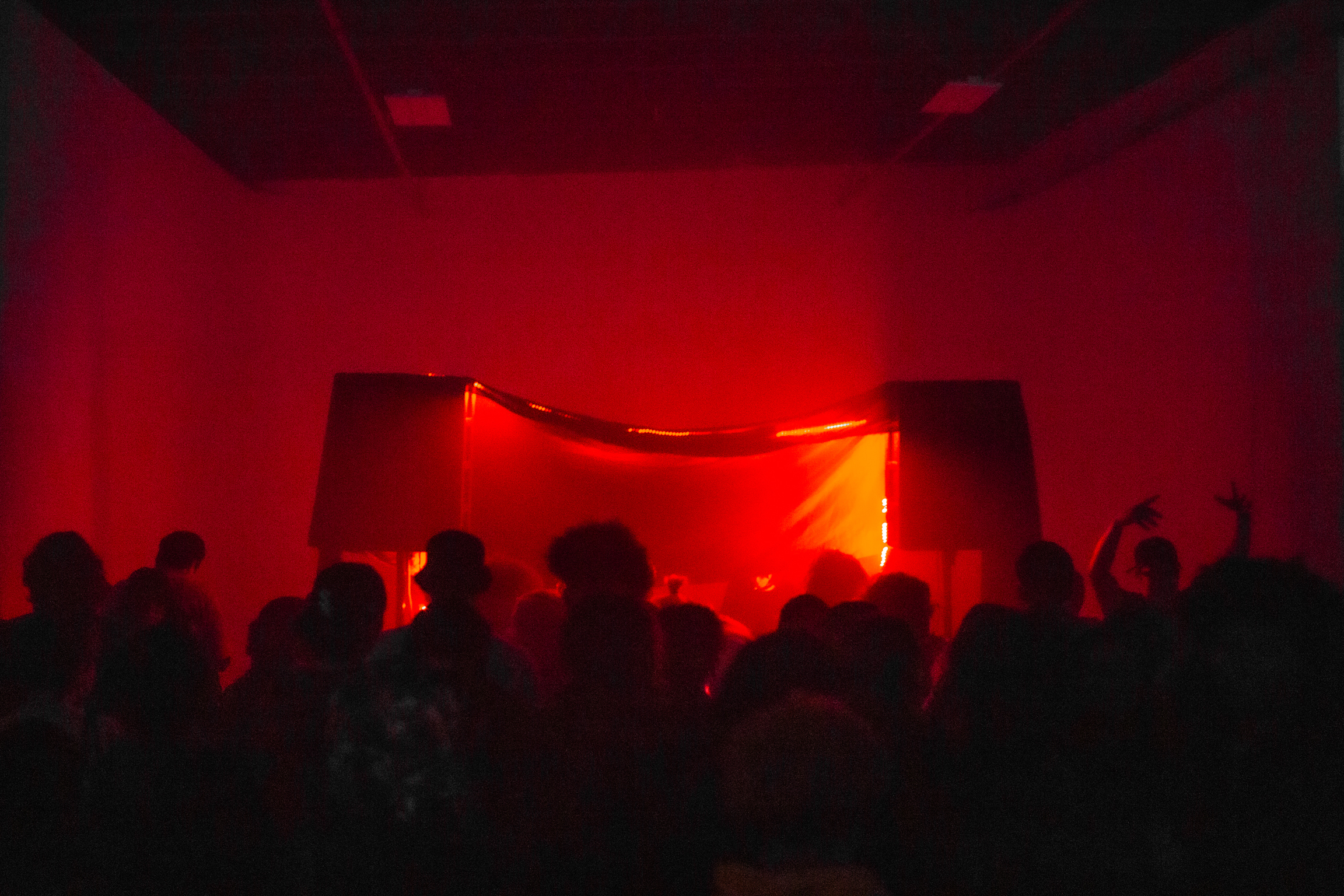 Crowd of silhouetted people in room bathed in deep red lighting with bright orange-red illuminated screen or stage area at centre.