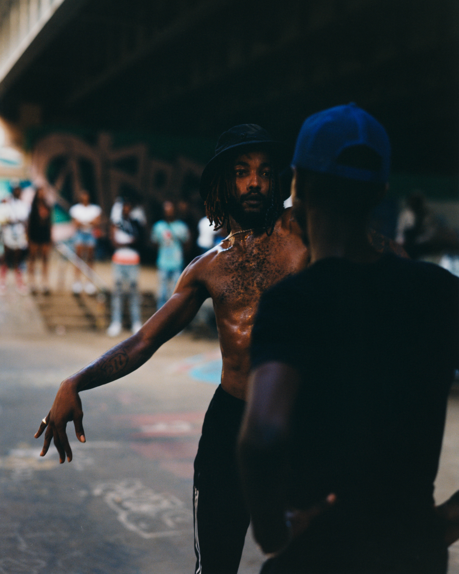 Man in blue cap and dark clothing dancing outdoors, with blurred crowd and building in background.