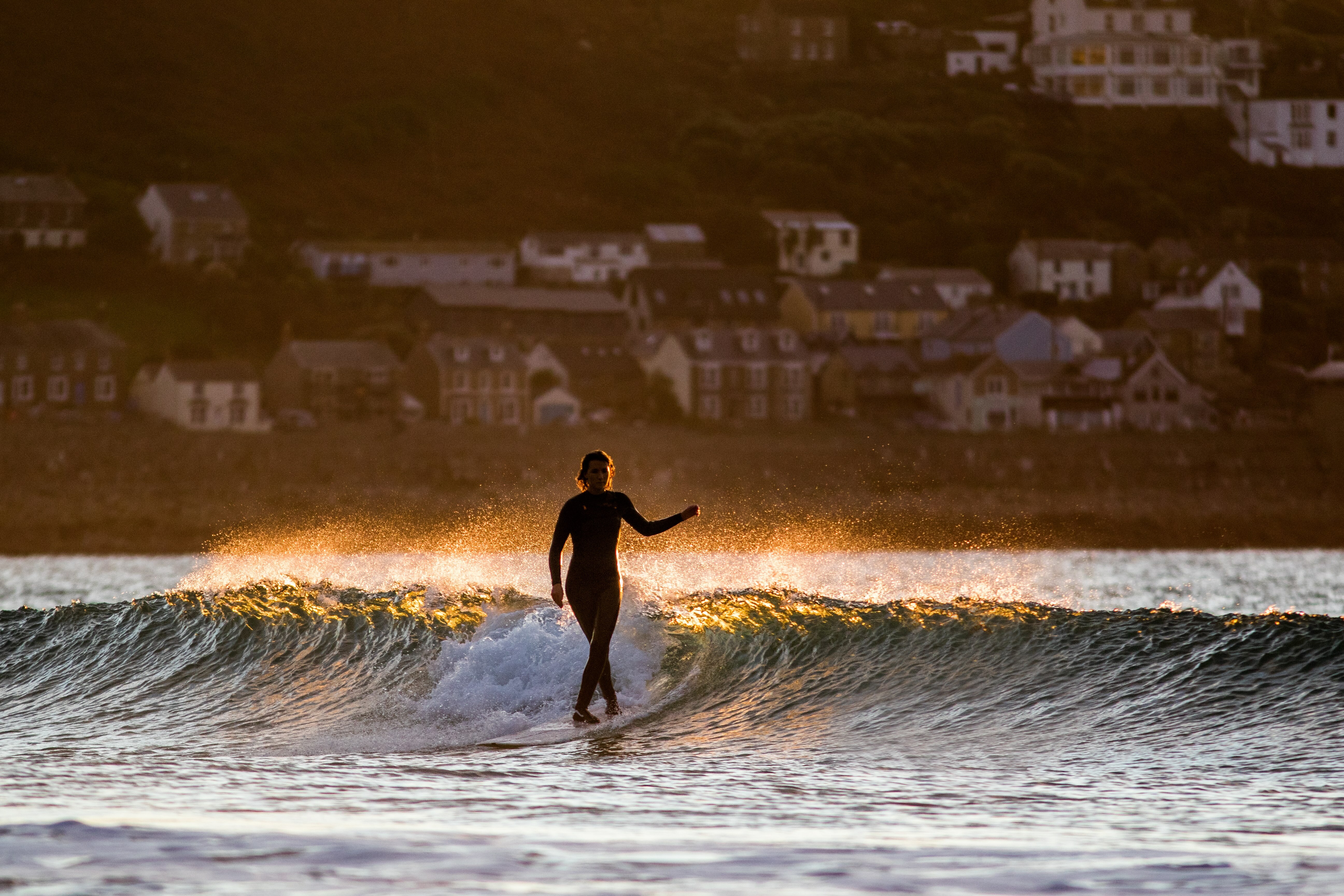 Silhouette of surfer riding large wave at sunset with lights of coastal town in background.