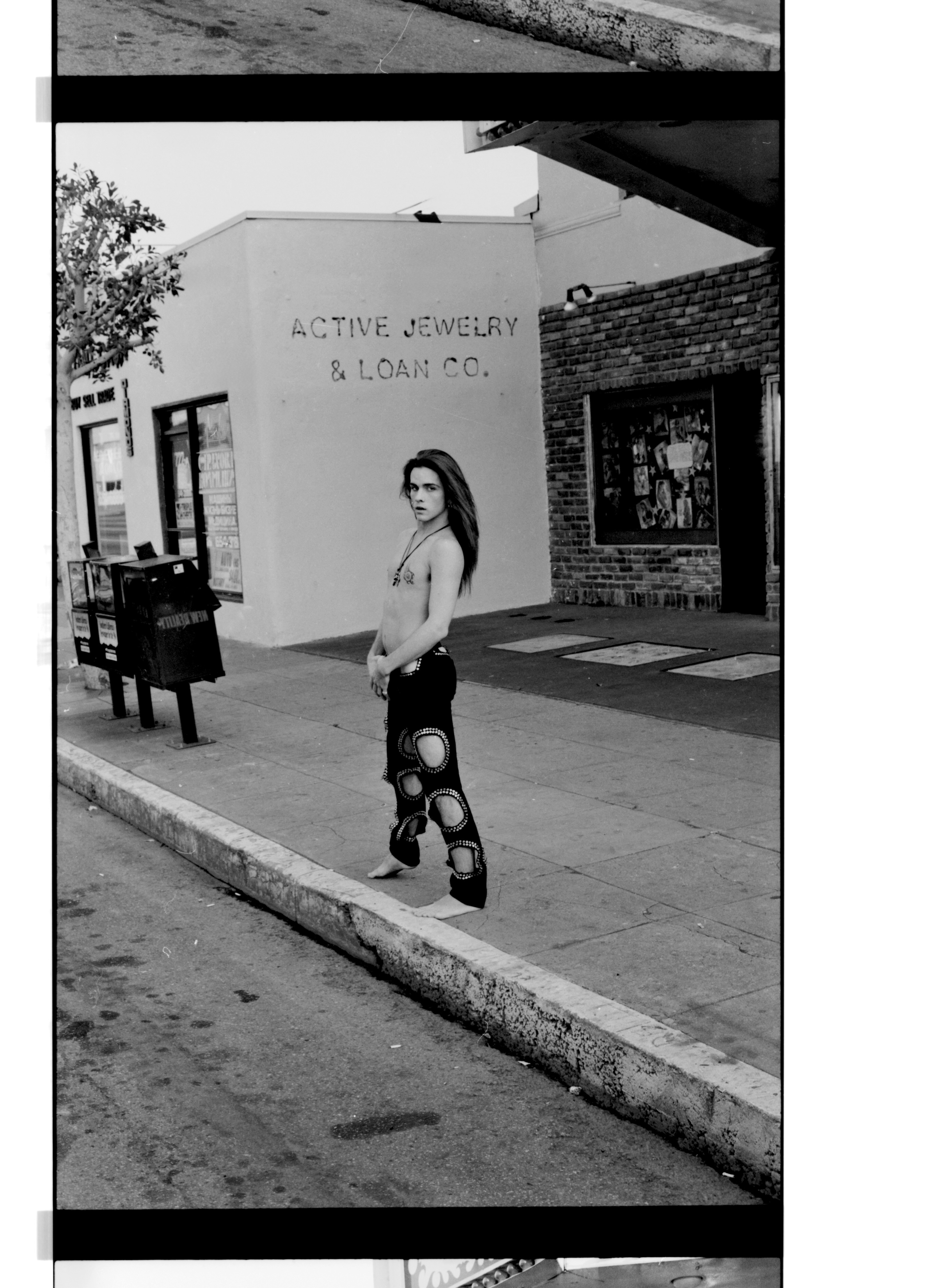 Black and white image of woman in patterned trousers walking on pavement outside "Active Jewelry & Loan Co." shopfront with brick facade.