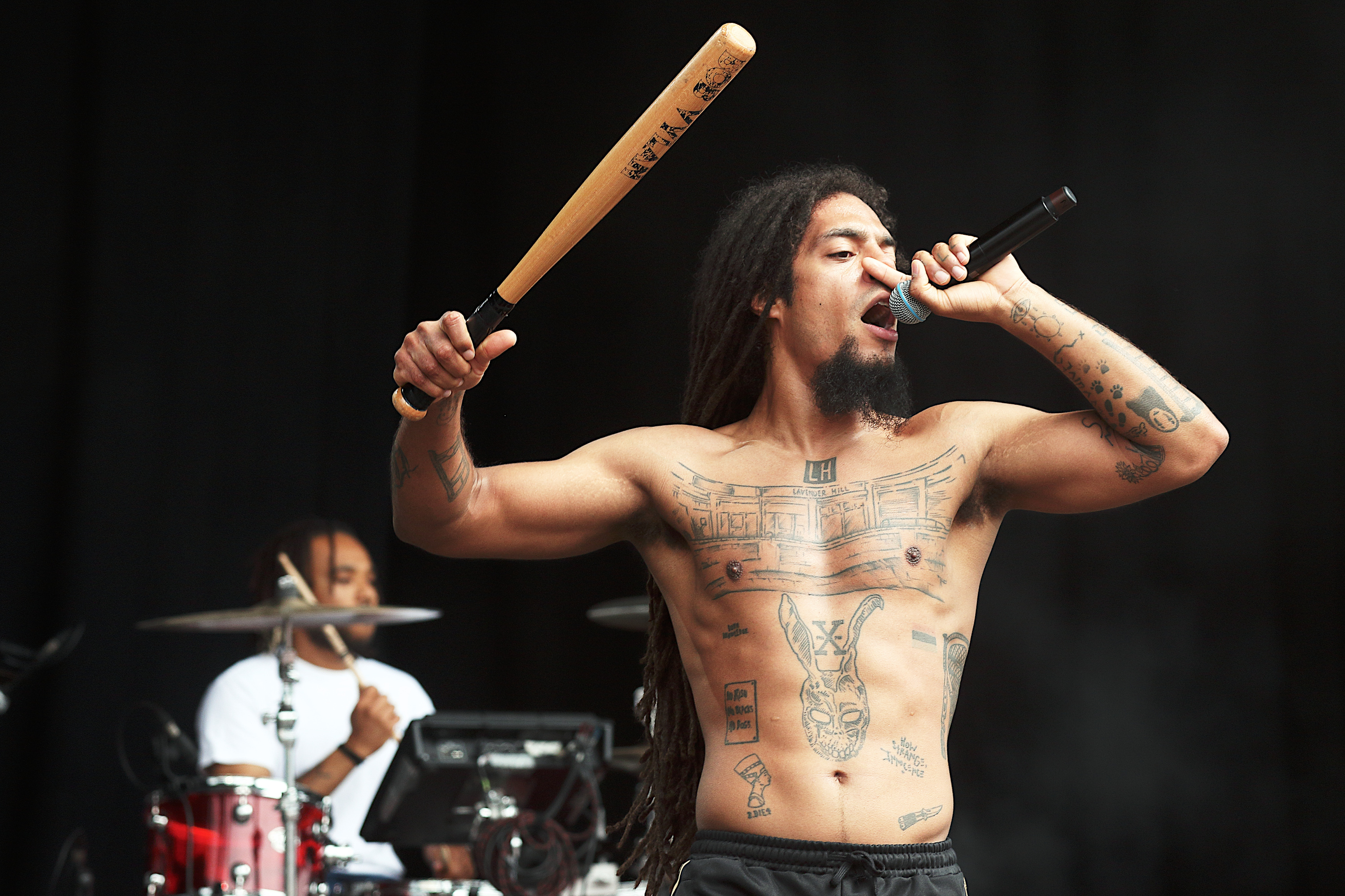 Shirtless male performer with long dark hair holds baseball bat aloft whilst singing into microphone, drummer visible in background.