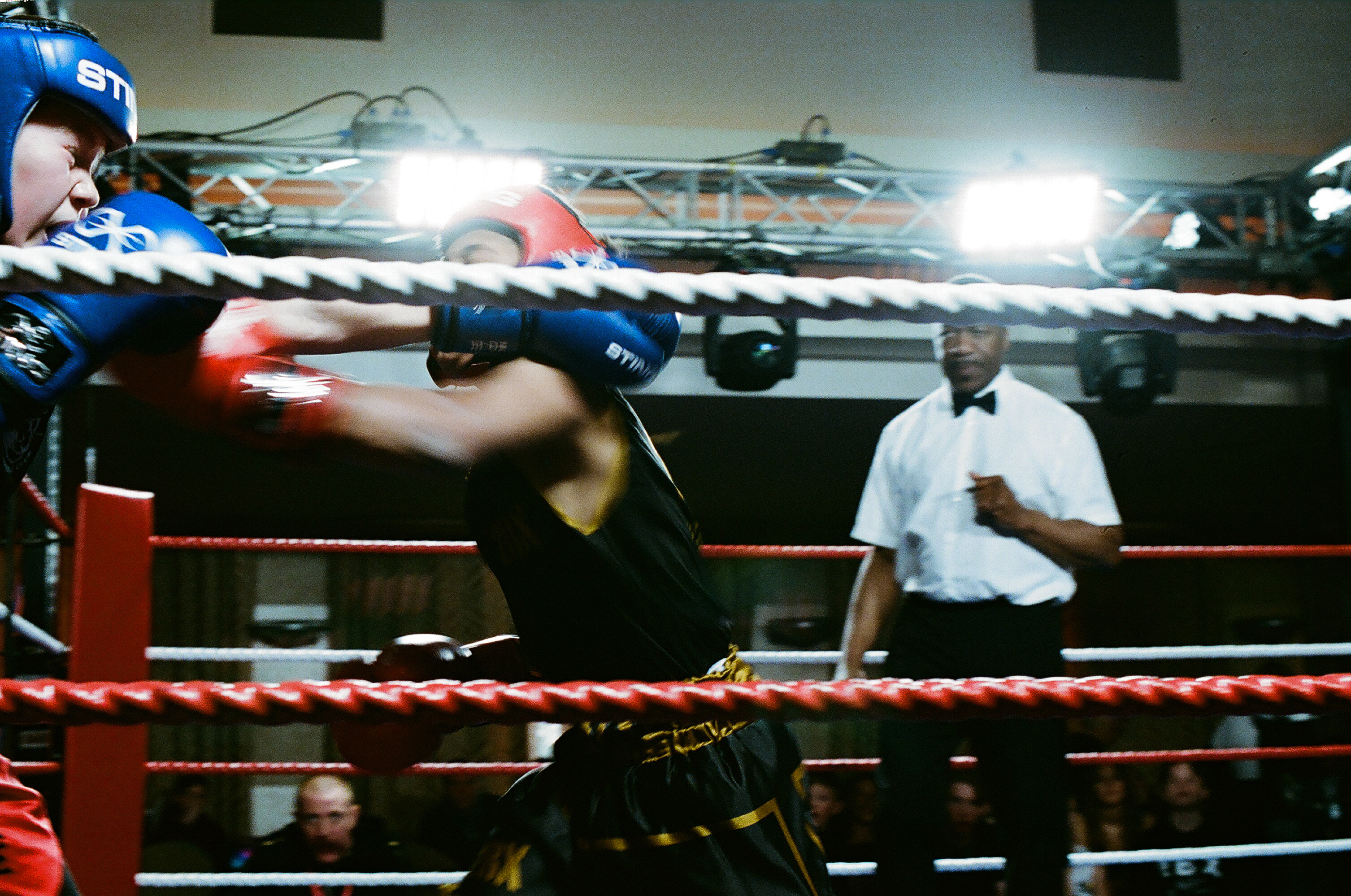 Two boxers in motion blur during match, referee in white shirt observing, red rope boxing ring with bright overhead lighting.