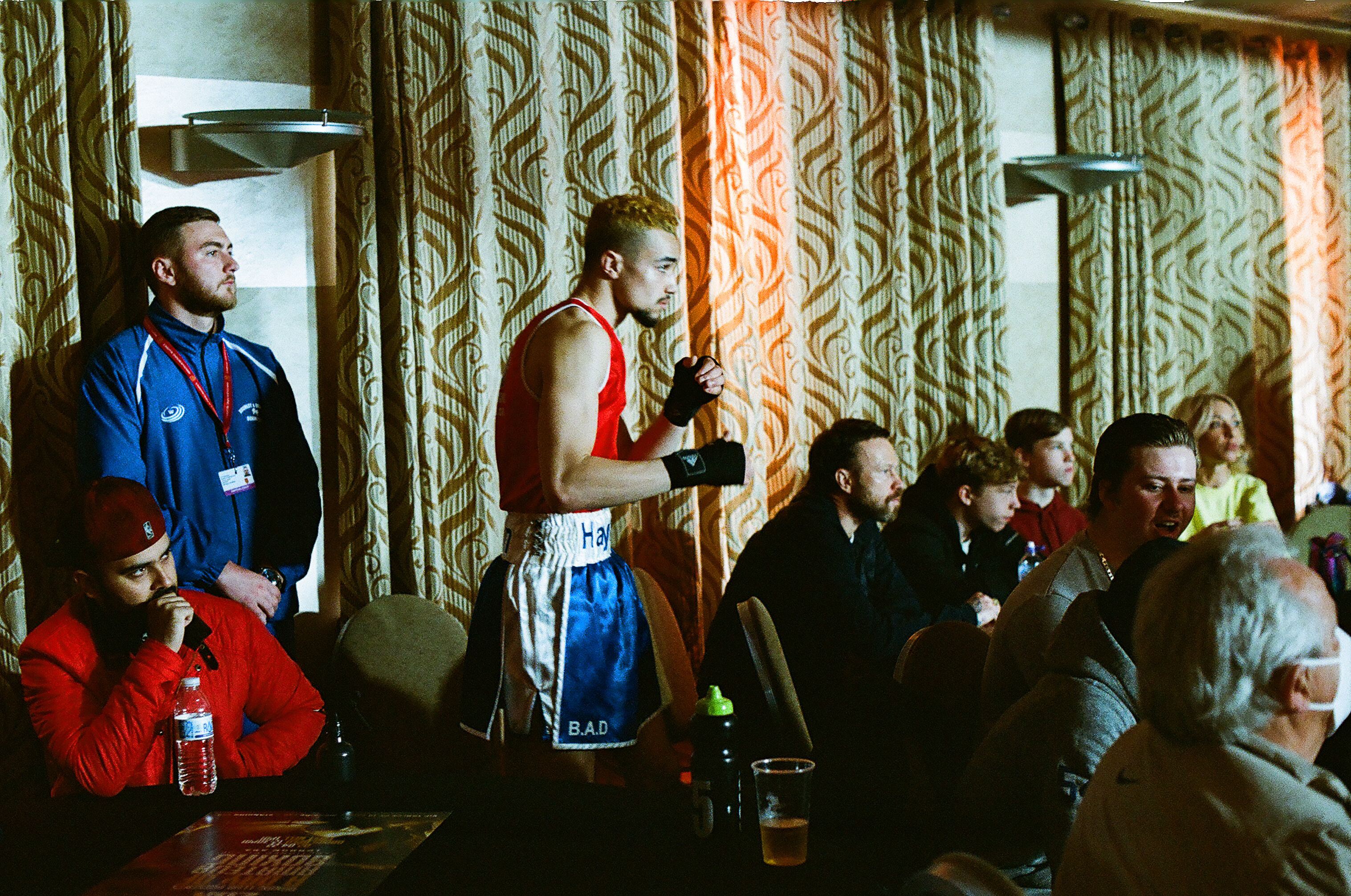 Basketball player in red vest and blue shorts holding trophy, standing amongst seated spectators in room with green patterned wallpaper.