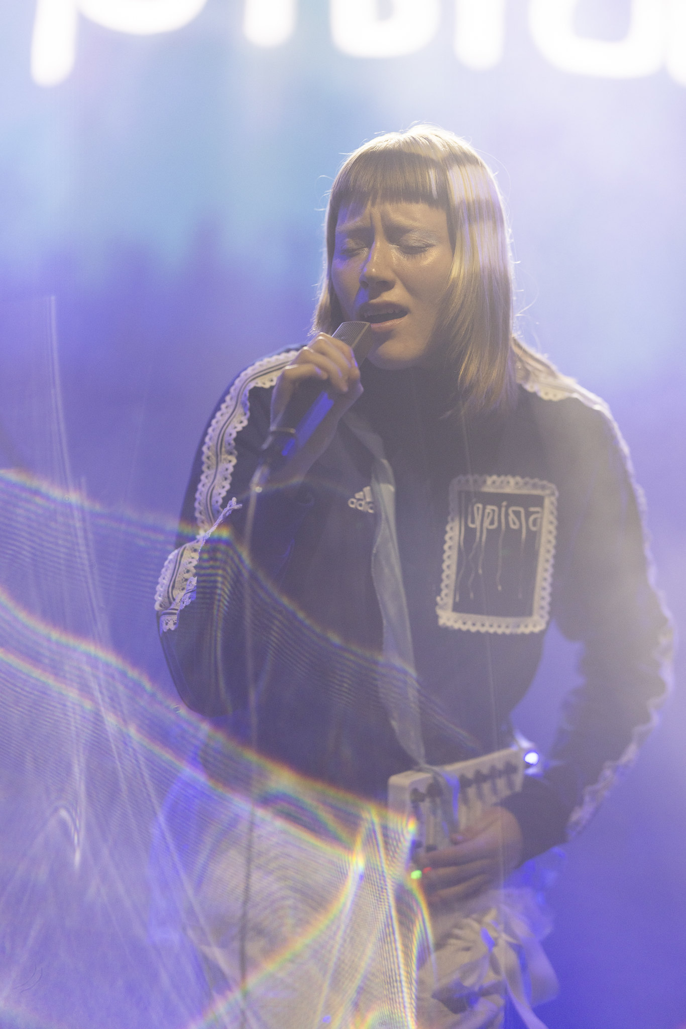 Woman with blonde bob singing into microphone on stage, wearing black jacket with silver patches, surrounded by purple and blue stage lighting.
