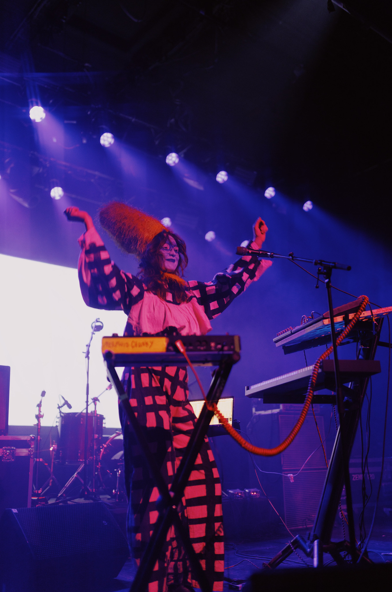 Performer on stage in red and black patterned costume with arms raised, surrounded by blue stage lights and musical equipment.