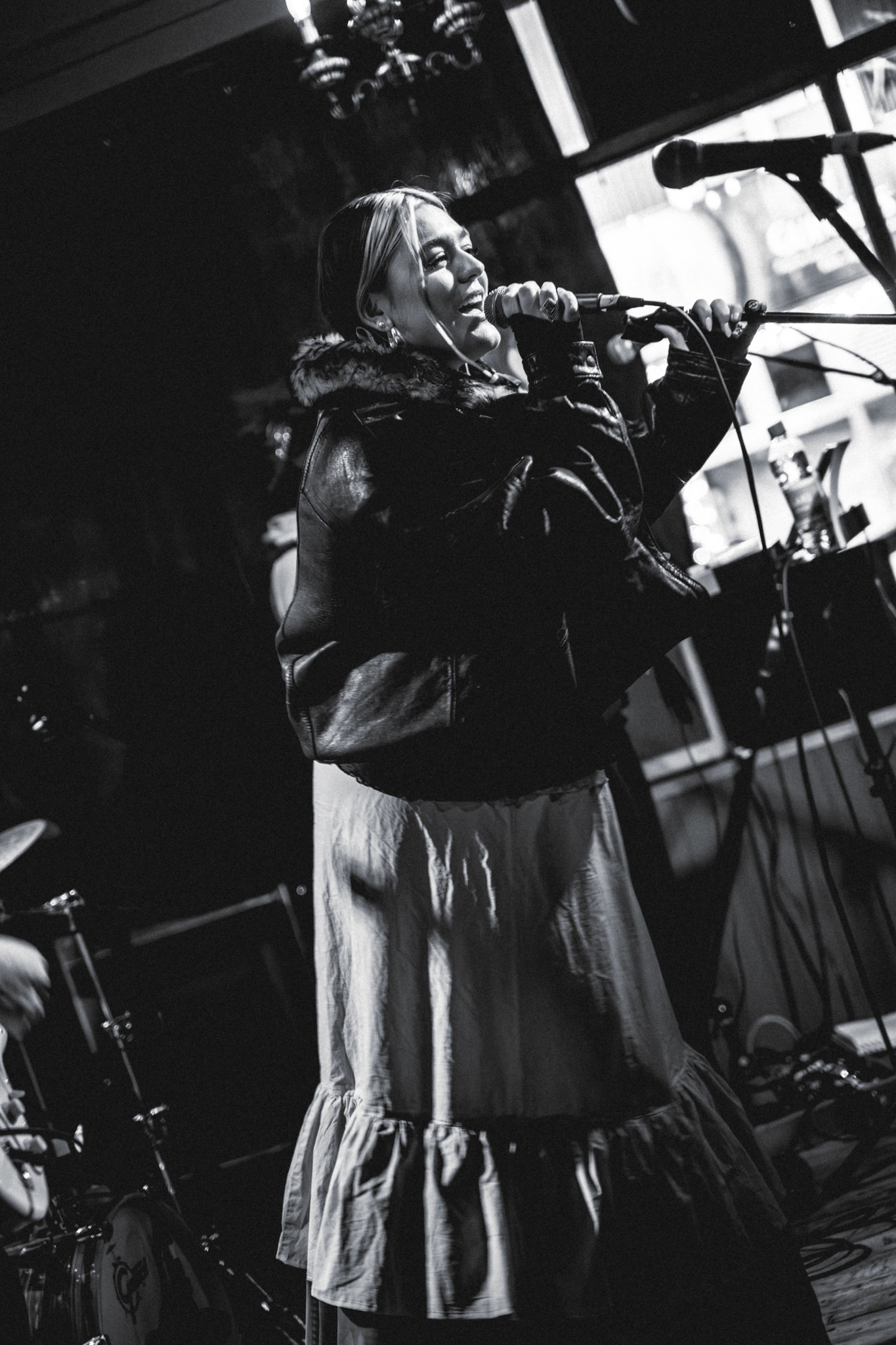 Woman in leather jacket singing into microphone on stage, black and white image with dramatic lighting and industrial venue backdrop.