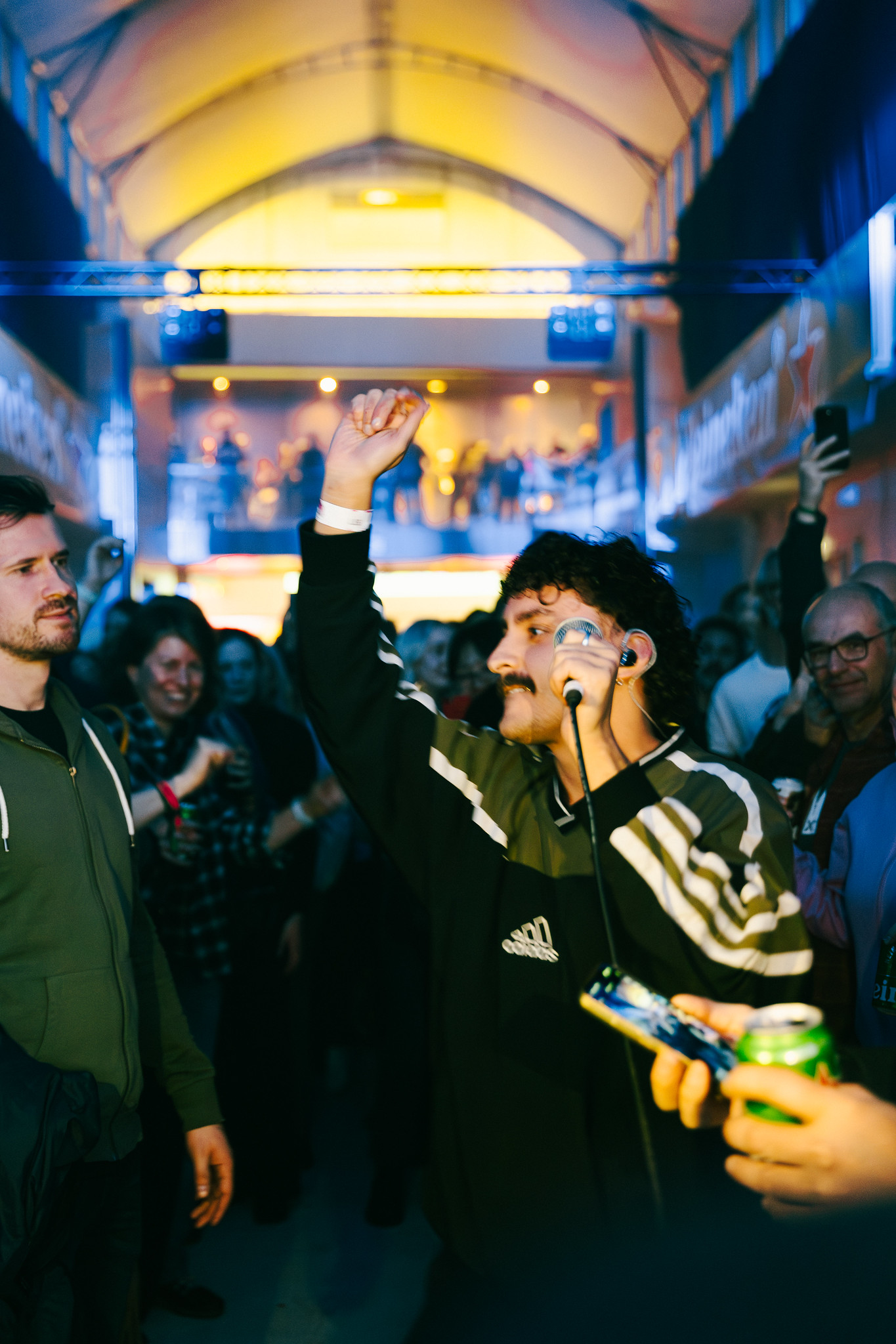 Man with microphone raising fist in crowded indoor venue with arched ceiling and warm yellow lighting.