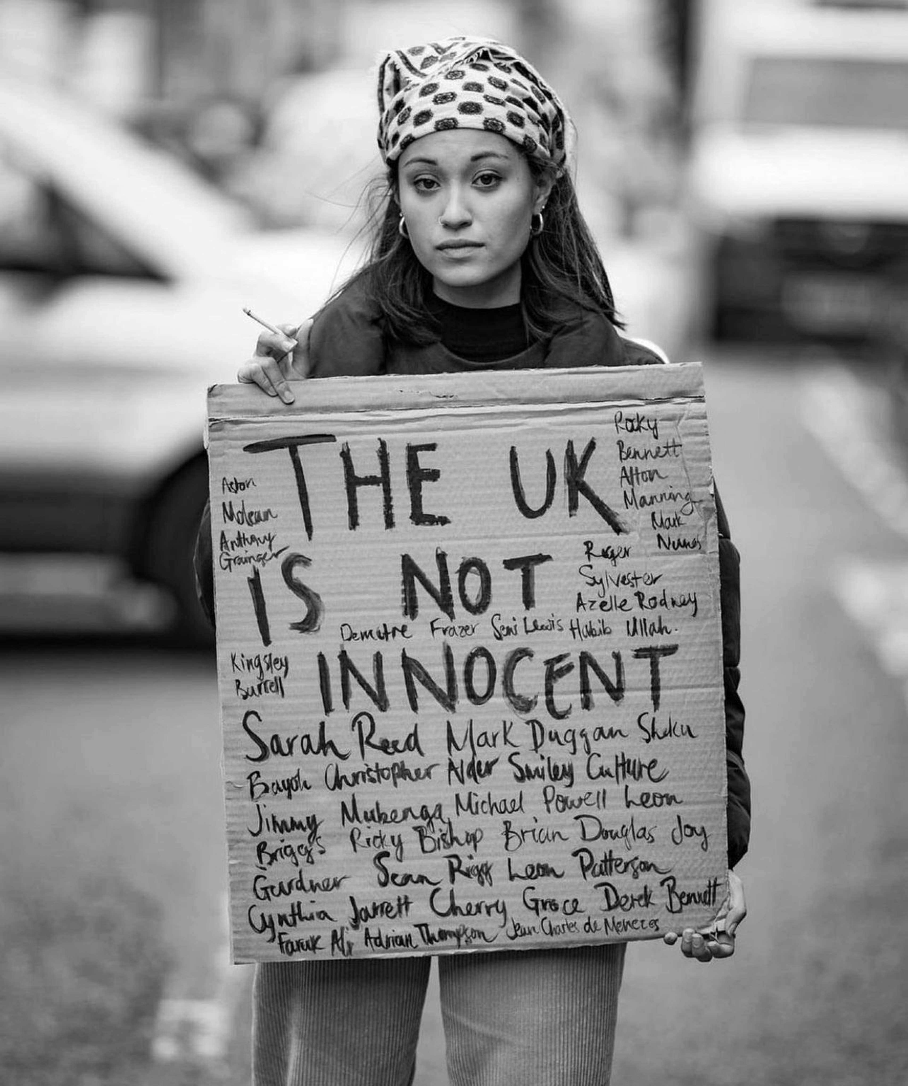 Young woman holding a sign reading "THE UK IS NOT INNOCENT" with various names written on it.