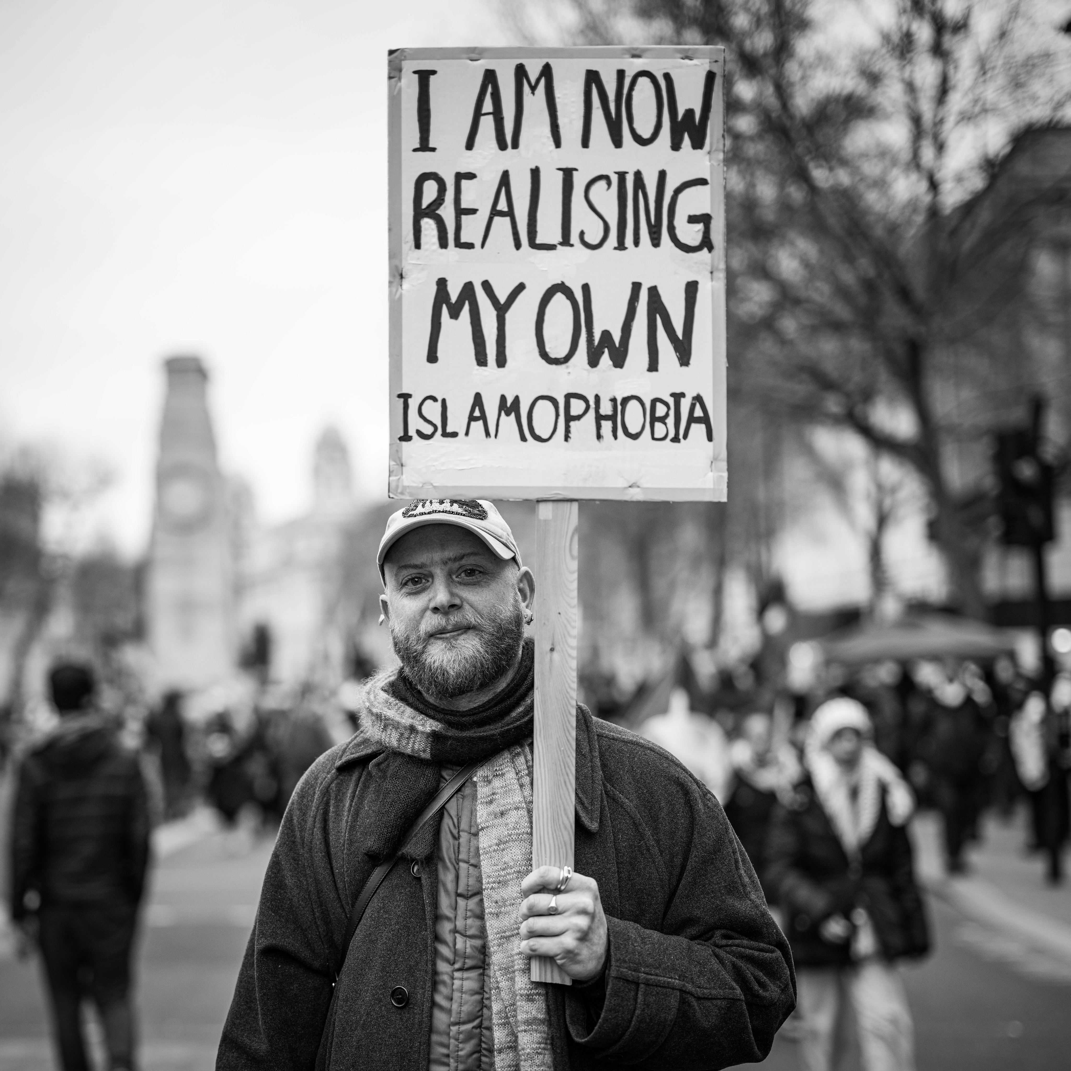Man holding sign reading "I AM NOW REALISING MY OWN ISLAMOPHOBIA" in black and white image.