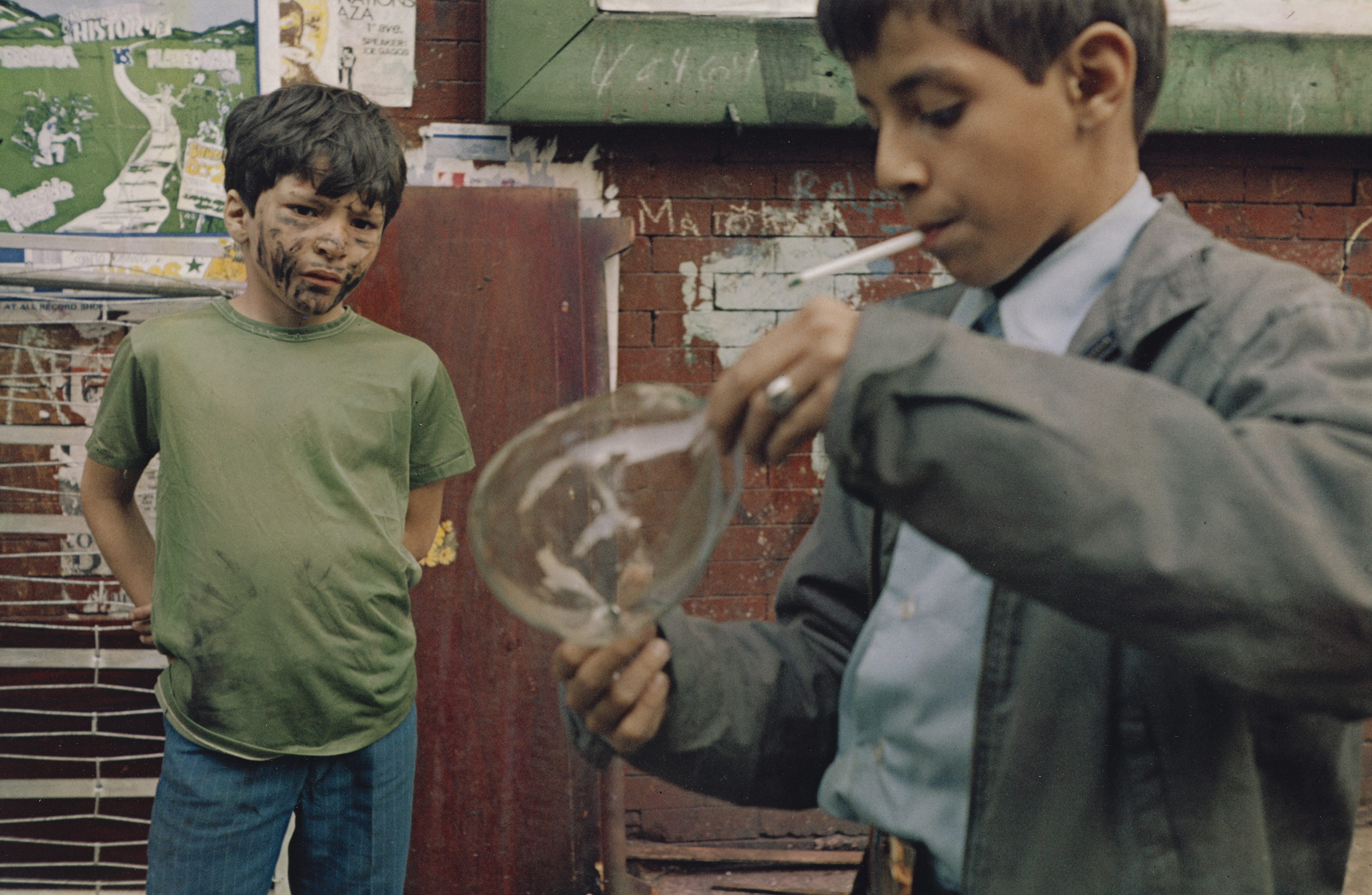 Two young boys, one wearing a green shirt and the other dressed formally, standing in front of a graffiti-covered wall and examining an object together.