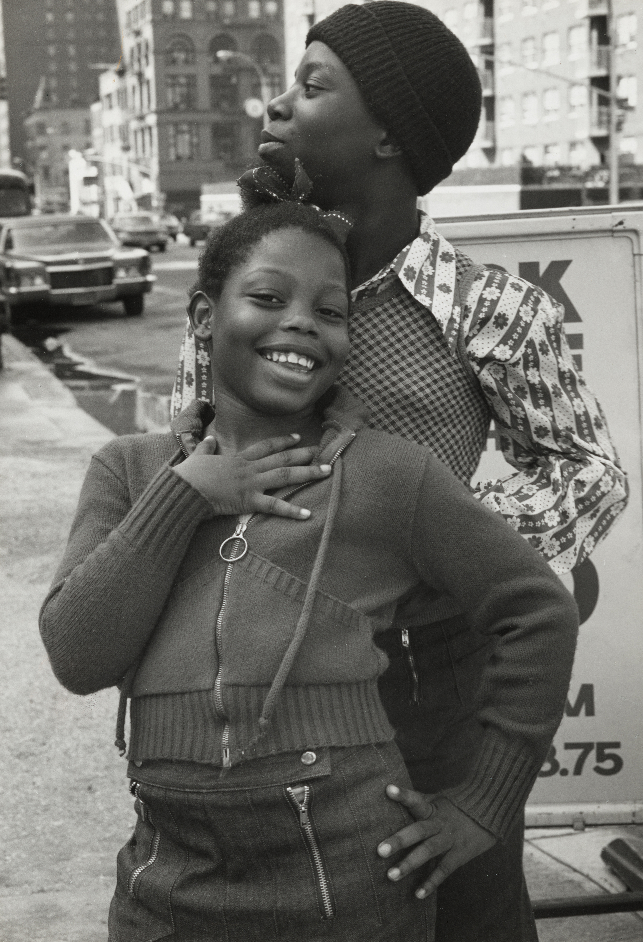 Two African-American women embracing, one smiling widely and the other looking tenderly at her companion. Black and white image of a city street in the background.