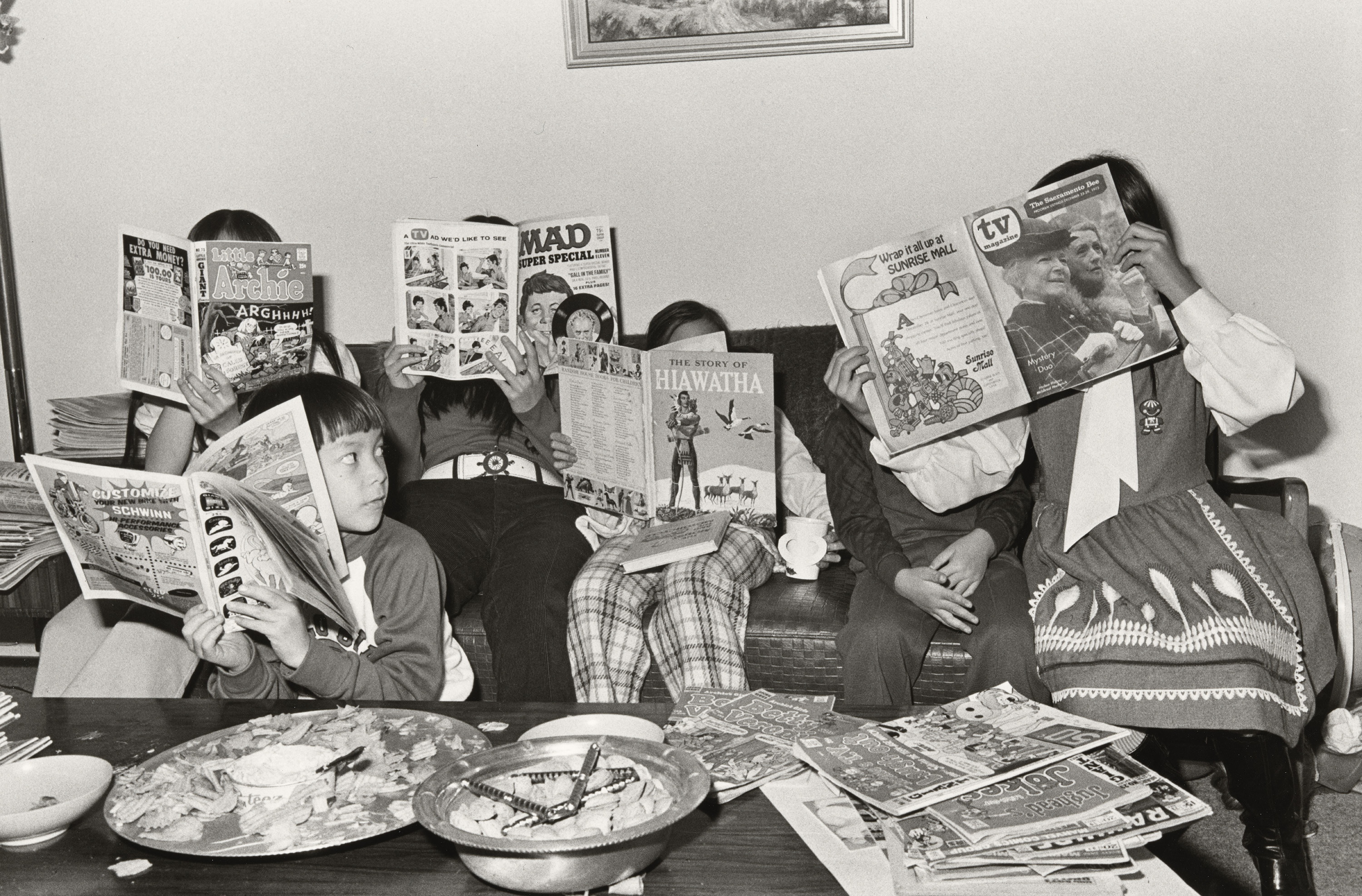 A group of people, likely children, sitting around a table surrounded by various comic books, magazines, and plates of food.
