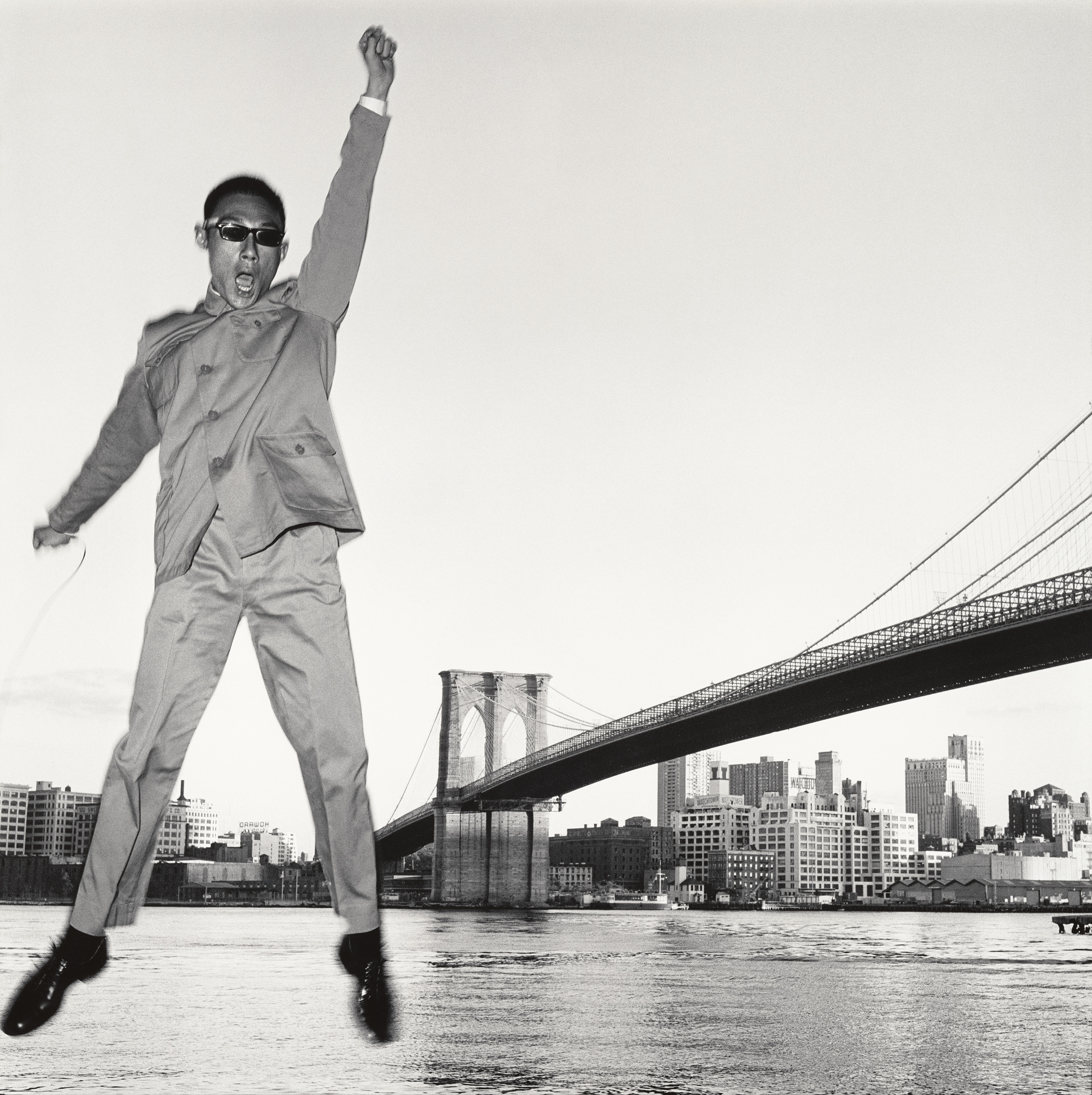 A person jumping with arms raised against a cityscape backdrop, including the Brooklyn Bridge.