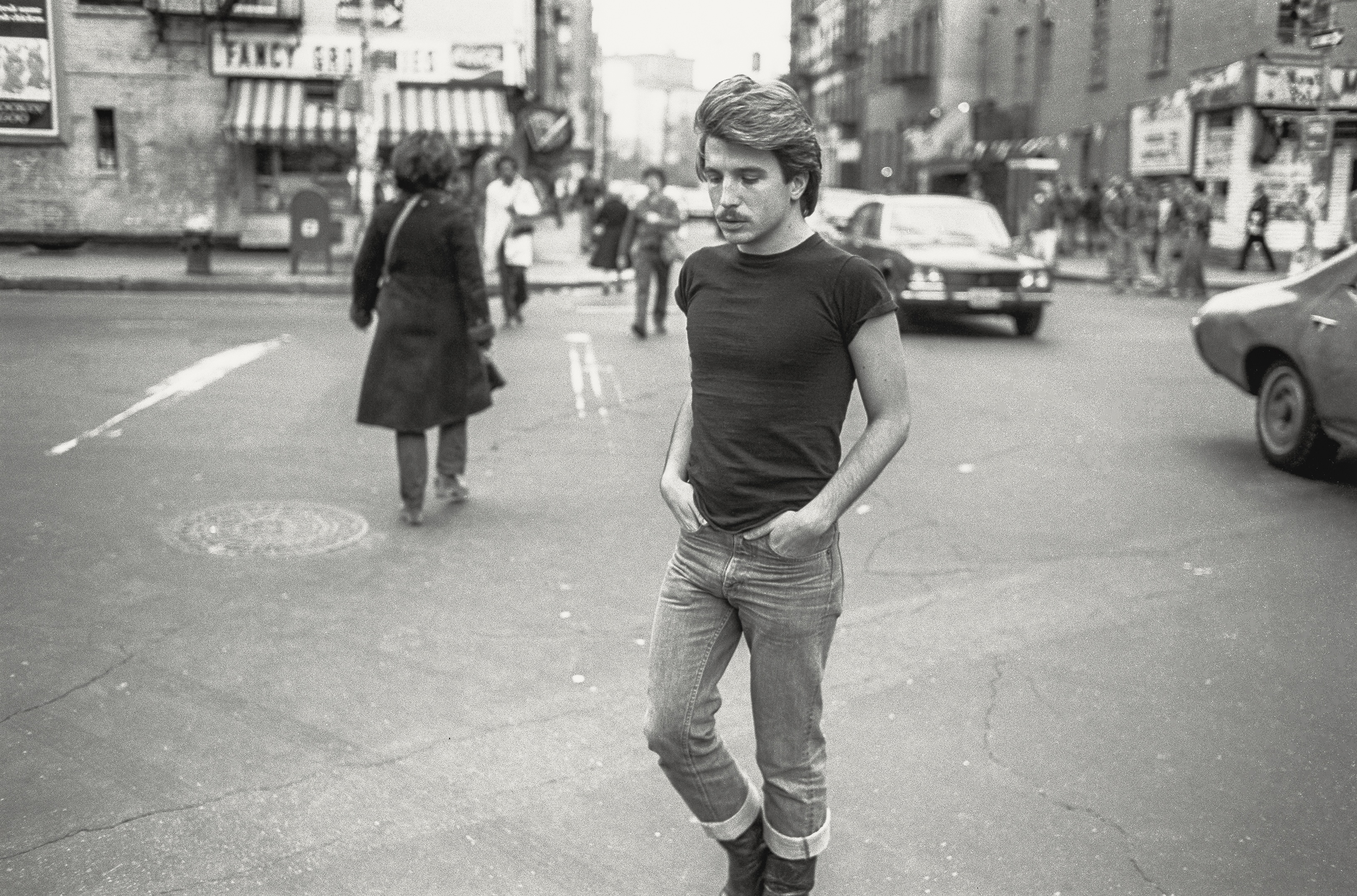 A young man stands on a city street, wearing a black top and blue jeans.