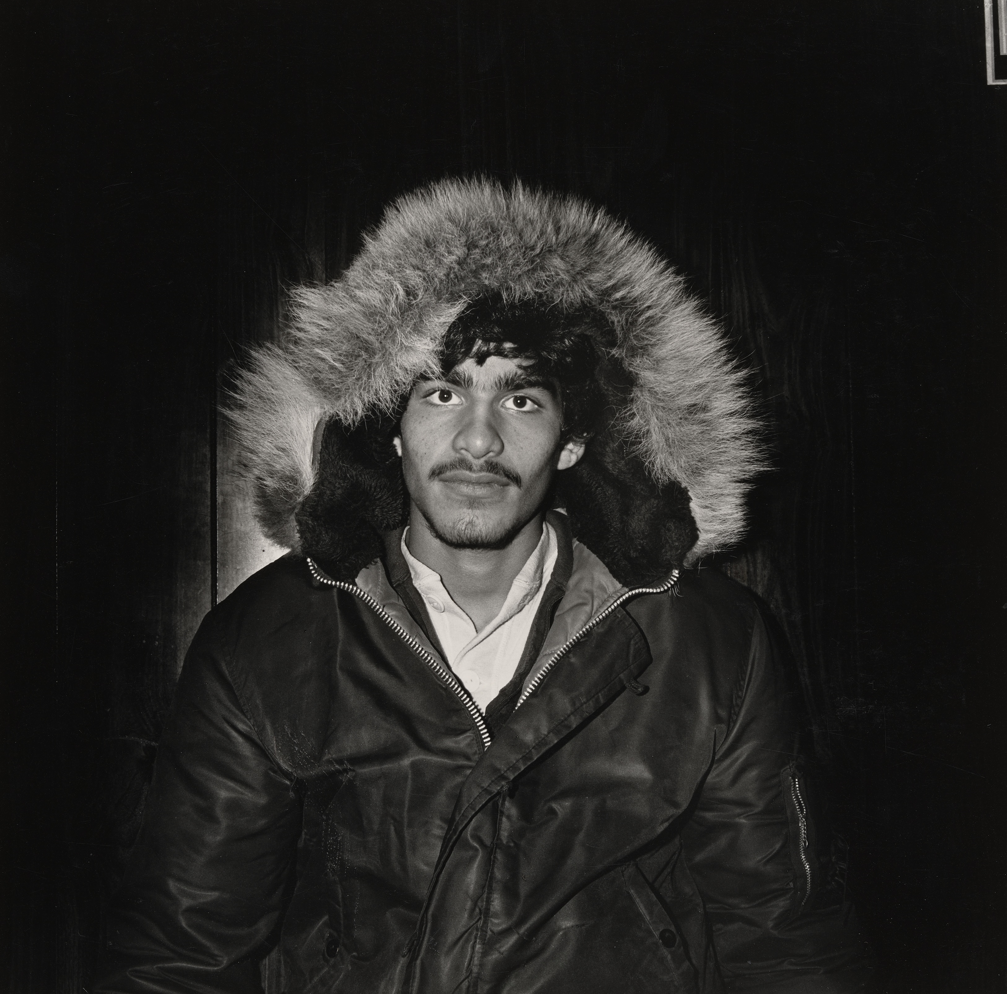 Individual wearing a hooded black jacket with fur trim, looking directly at the camera against a dark background.