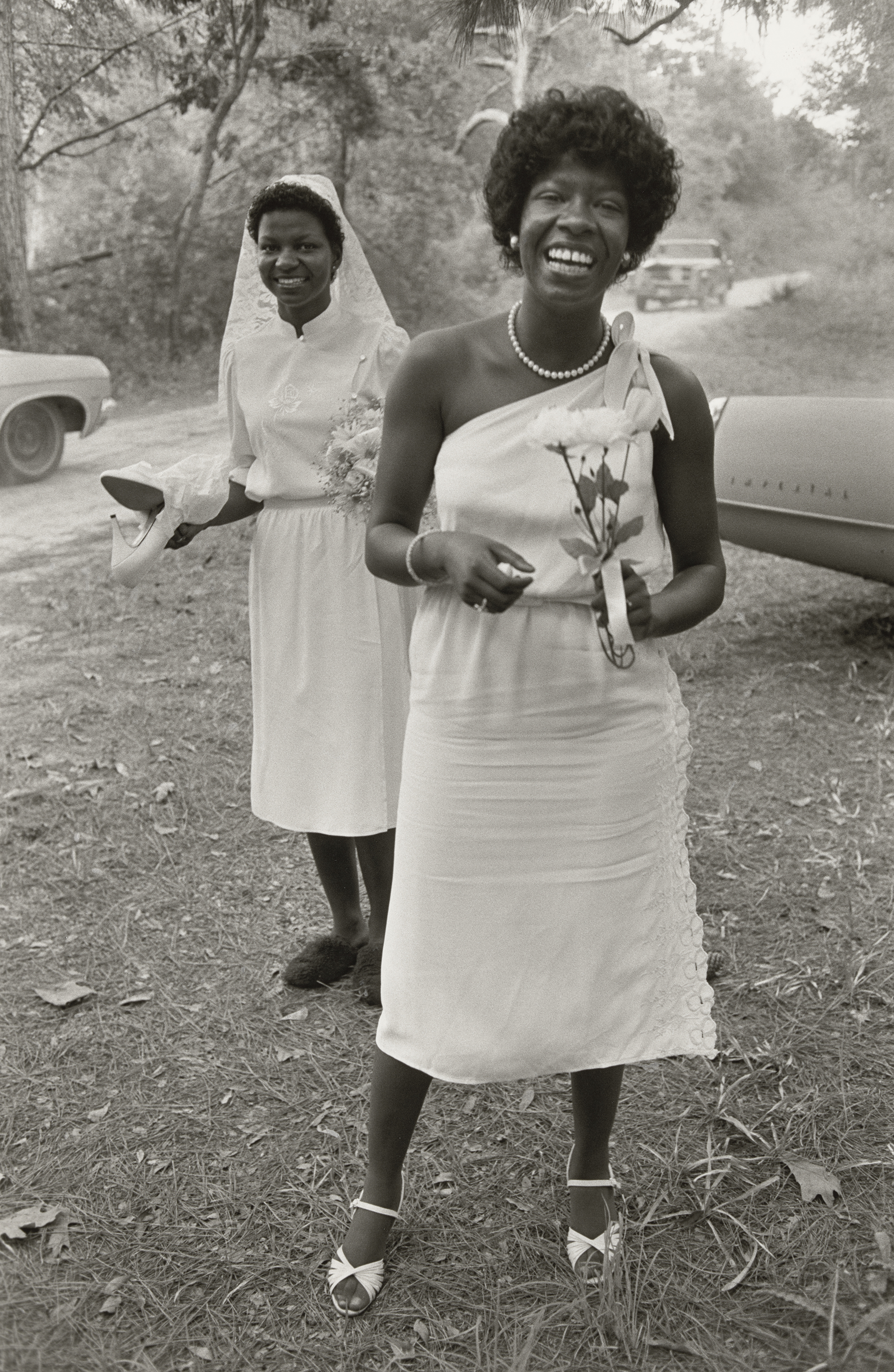 Two smiling women wearing white dresses and standing in a wooded area.