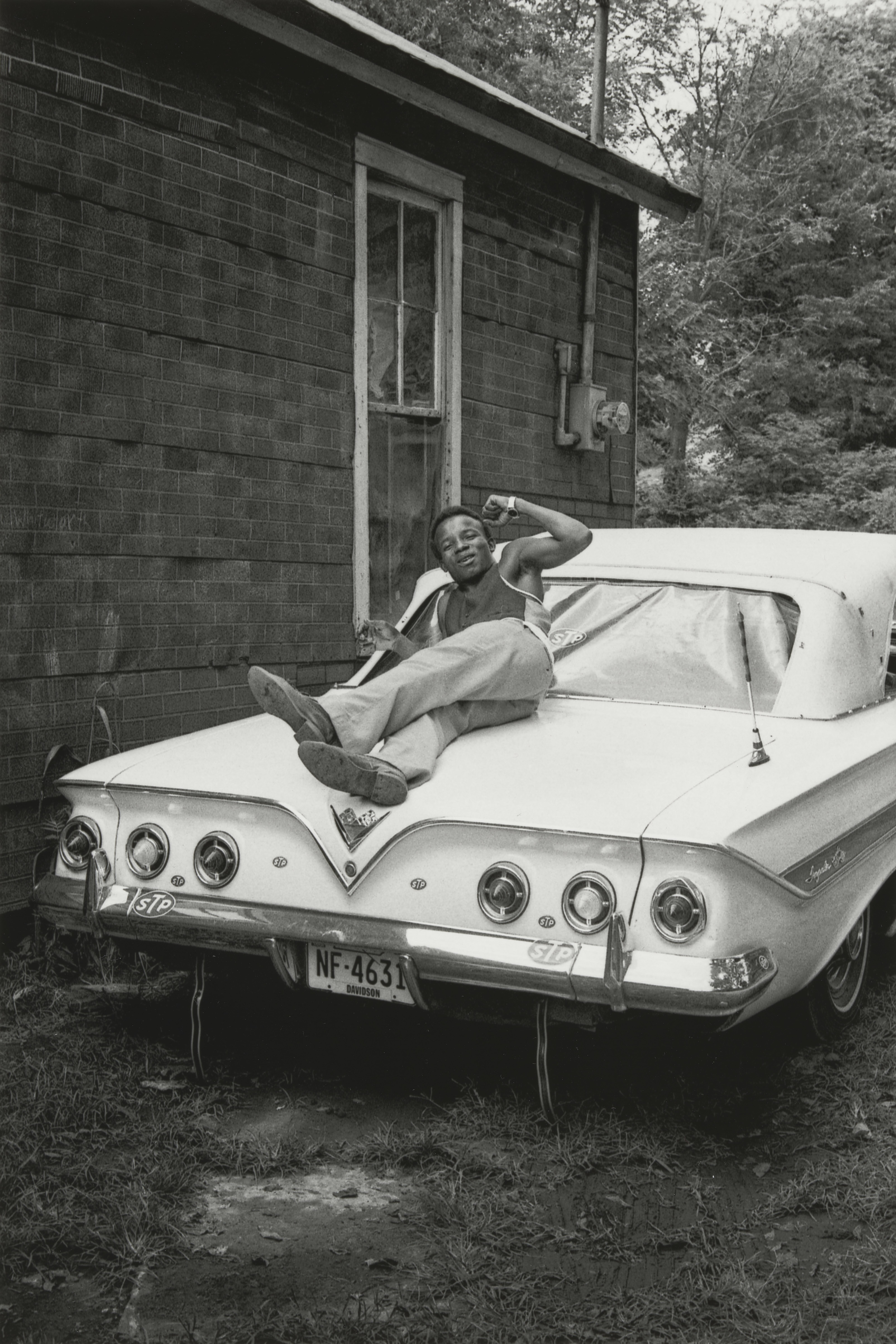 Black and white photograph of a man reclining on the back of a vintage white sports car parked in a grassy area with a brick building in the background.