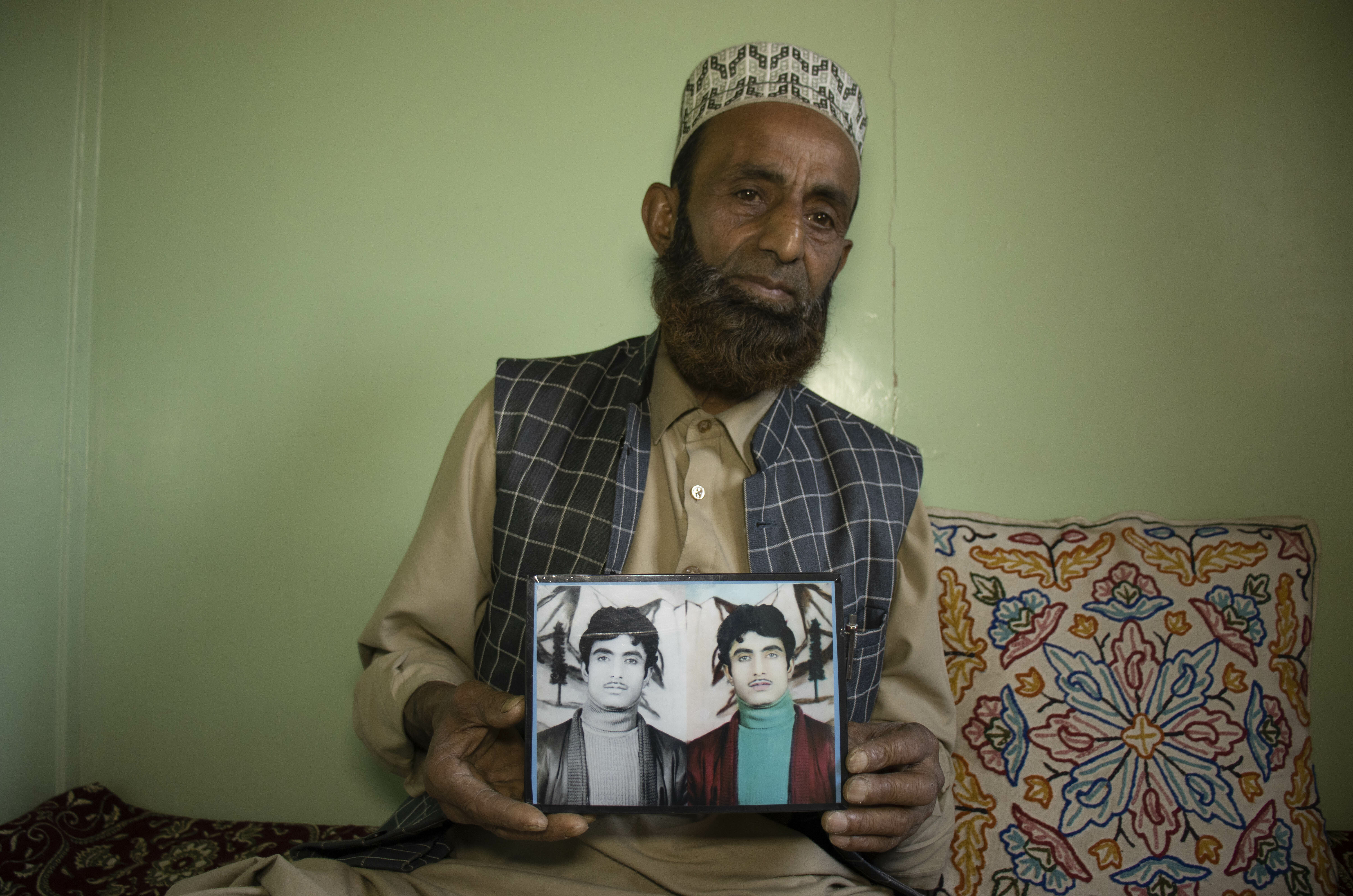 Bearded man in traditional attire holding a framed photograph of two young men.