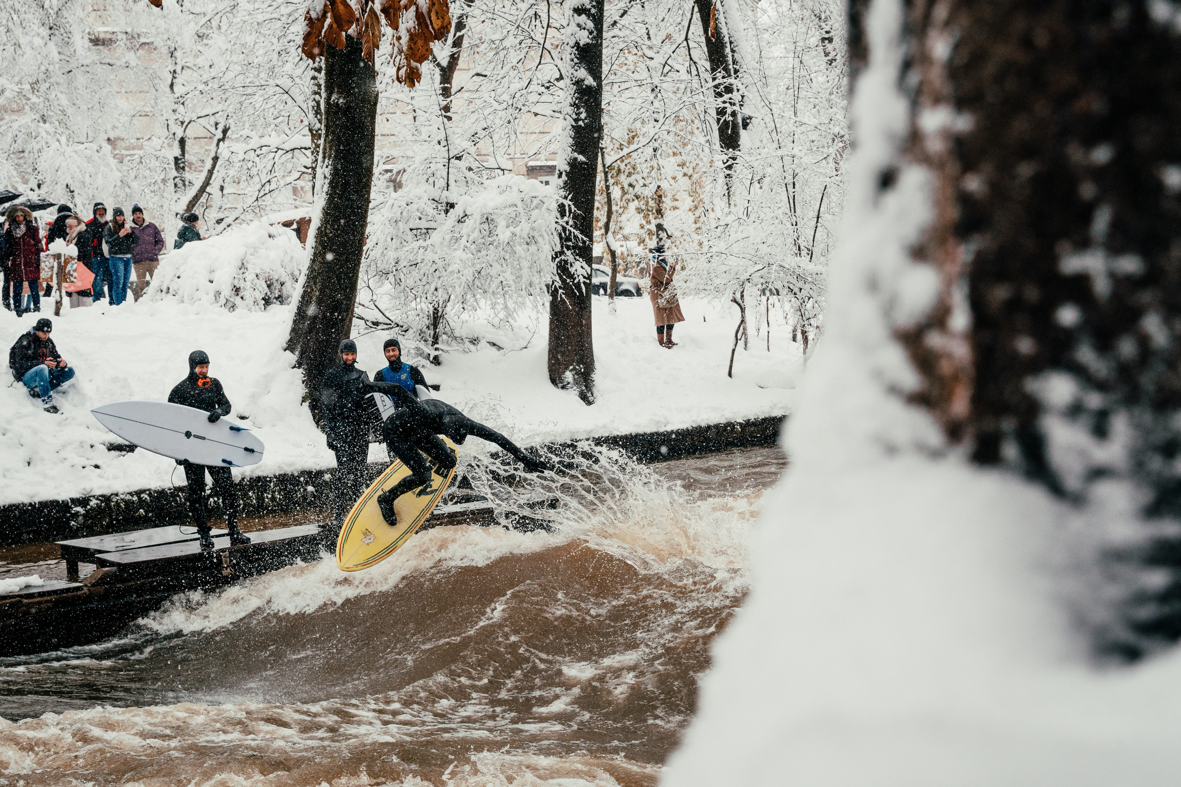 Person surfing on brown water stream flowing through snowy landscape with spectators watching from white banks amongst bare trees.