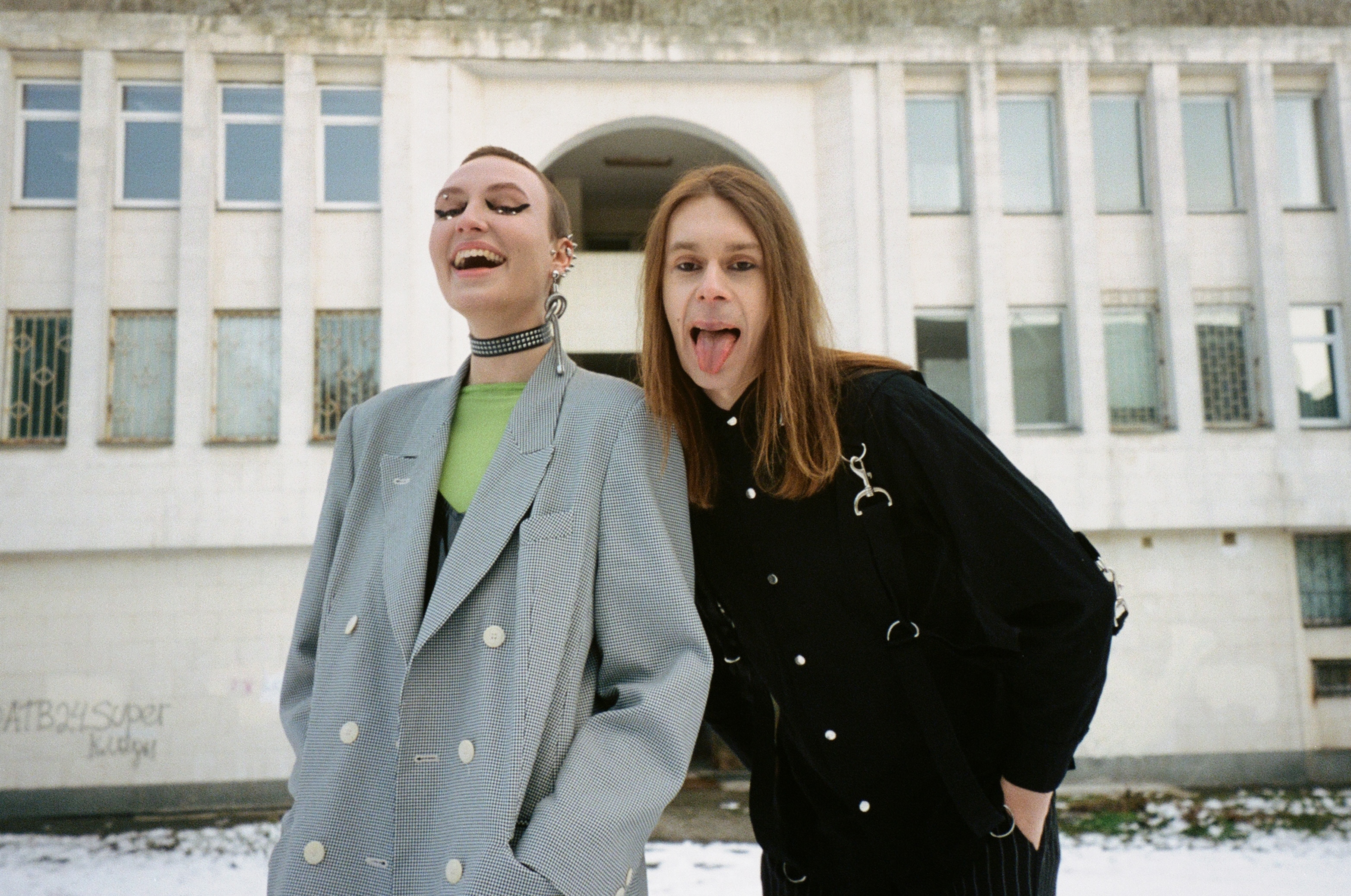 Two young women making silly facial expressions, one with a grey coat and the other in a black coat, in front of a white building.