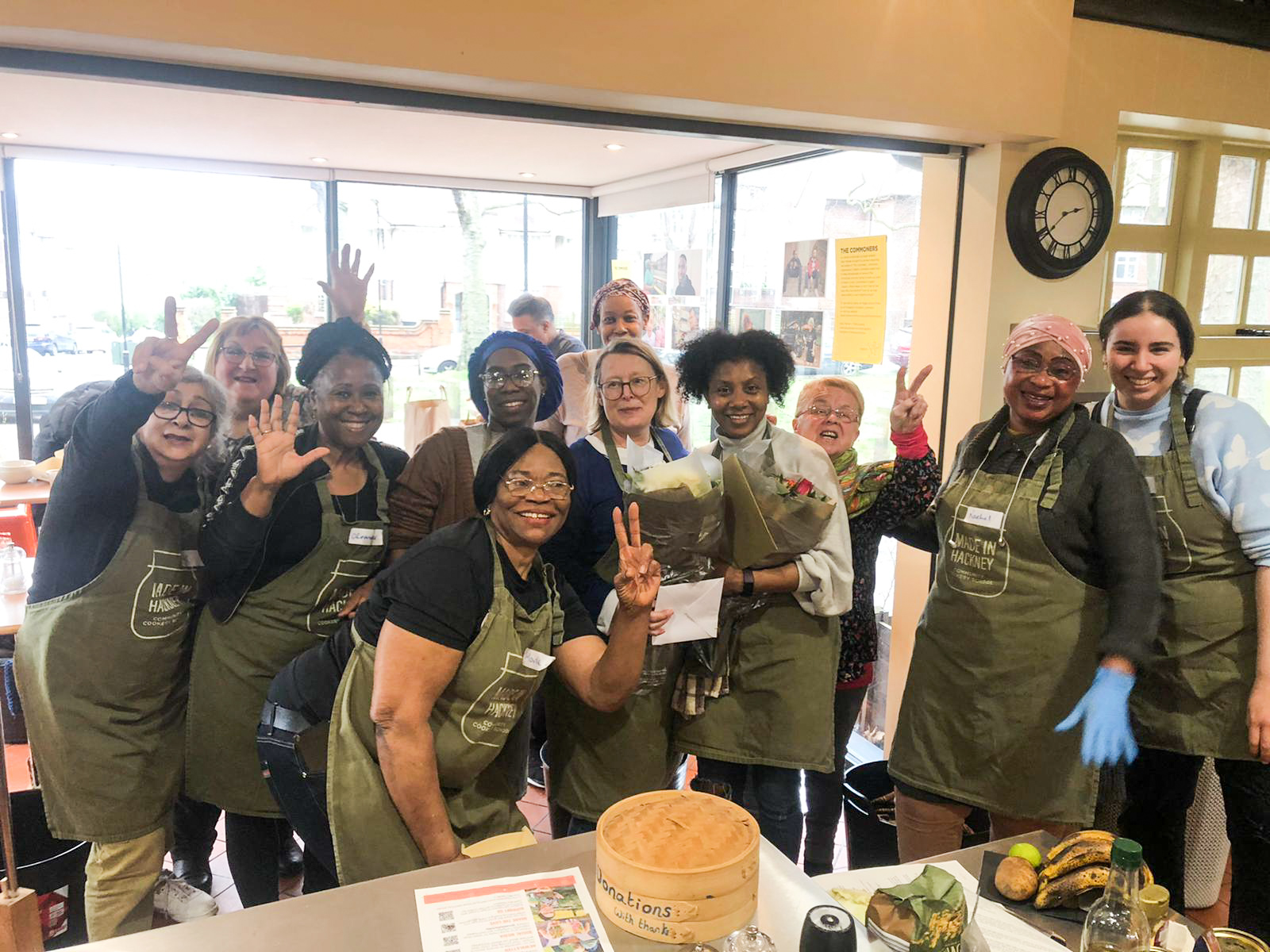 Group of people in green aprons waving at camera in bright room with large windows, clock on wall, and food items on table.