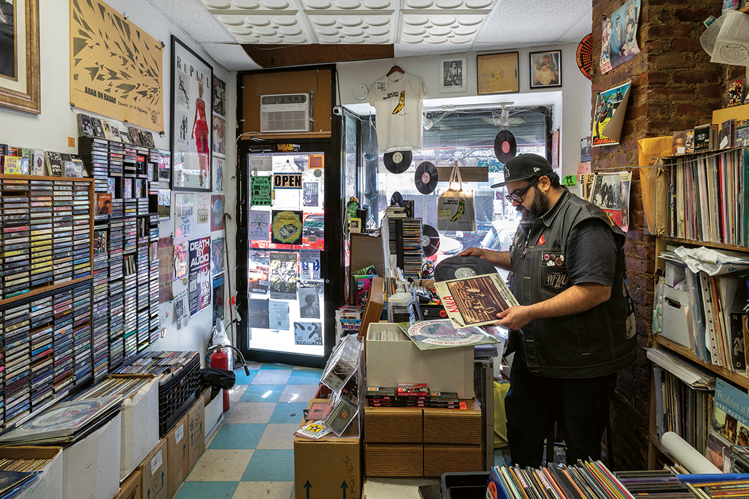 Man in dark clothing examining vinyl record in cluttered record shop with wall-mounted displays and shelves of albums