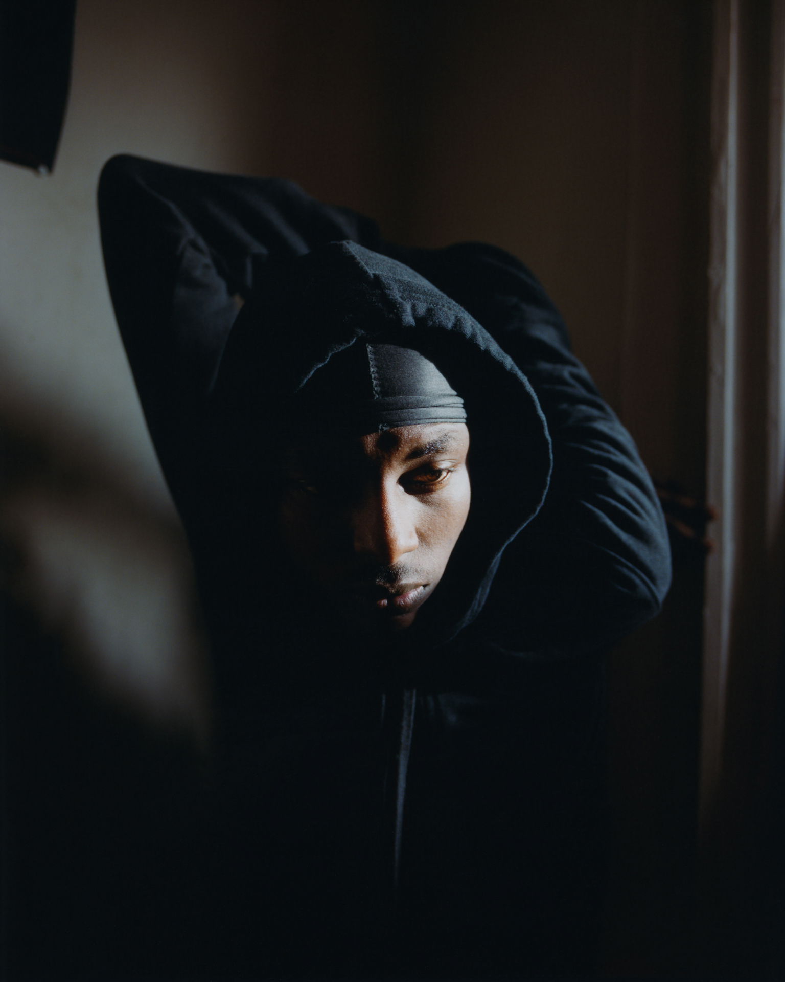 Person in dark hooded jacket with hood up, face partially visible in shadow, indoor setting with warm lighting.