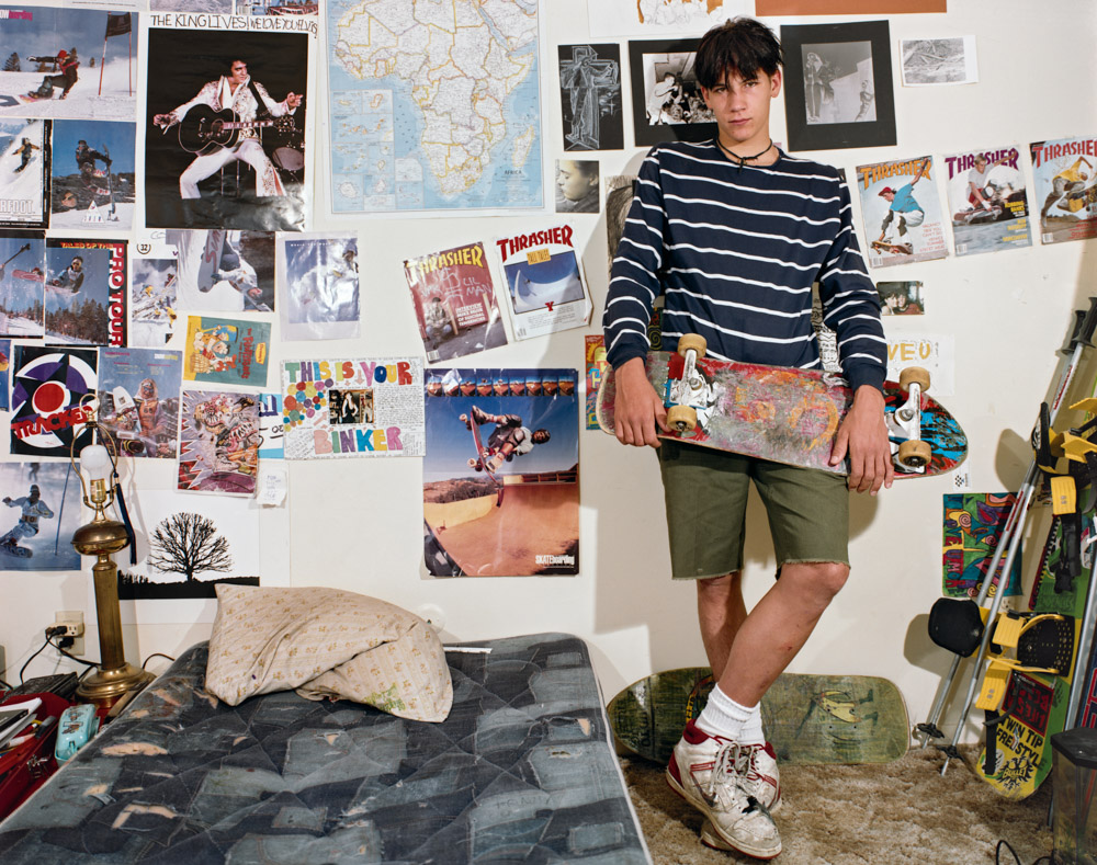 Young man in navy striped jumper and olive shorts stands in bedroom with walls covered in skateboarding posters and magazine clippings.