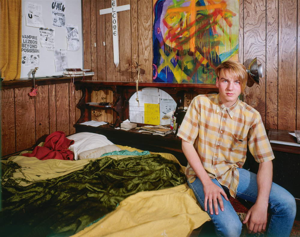 Young man in checked shirt sits on bed in wood-panelled room with colourful artwork, papers, and sleeping bags.