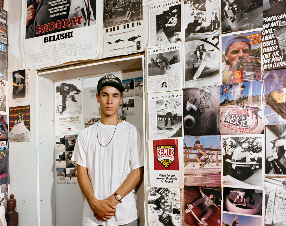 Young man in white t-shirt and black cap standing against wall covered in skateboarding magazine pages and posters.