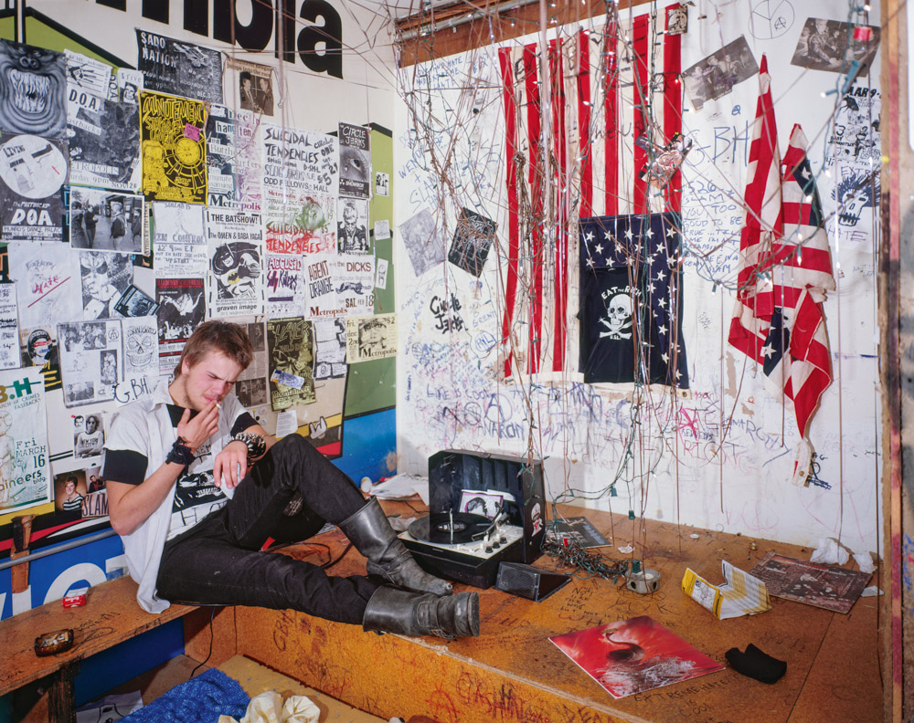 Young man sitting on floor in corner of room covered with posters, stickers, and graffiti. Red and white striped flag on wall.