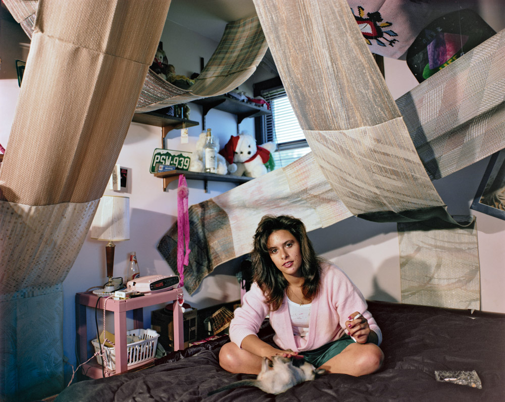 Woman in pink pyjamas sits cross-legged in makeshift fort constructed from white sheets and furniture in domestic interior.