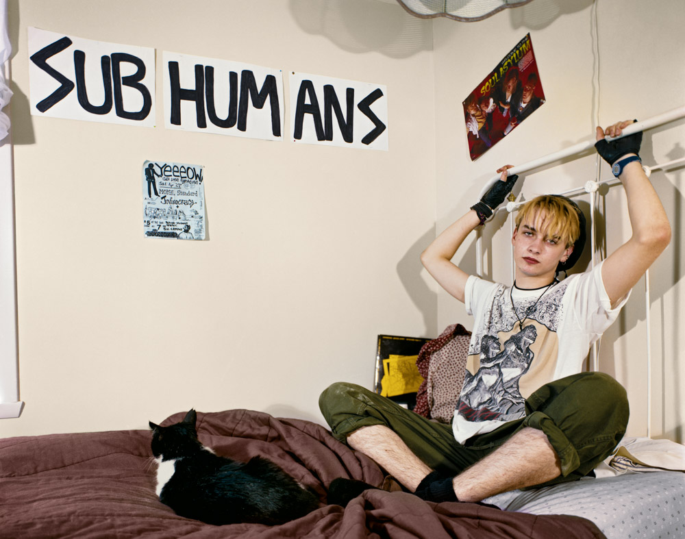 Person with blonde hair sits cross-legged on bed, arms raised, wearing grey t-shirt and green trousers. "SUB HUMANS" painted on wall.