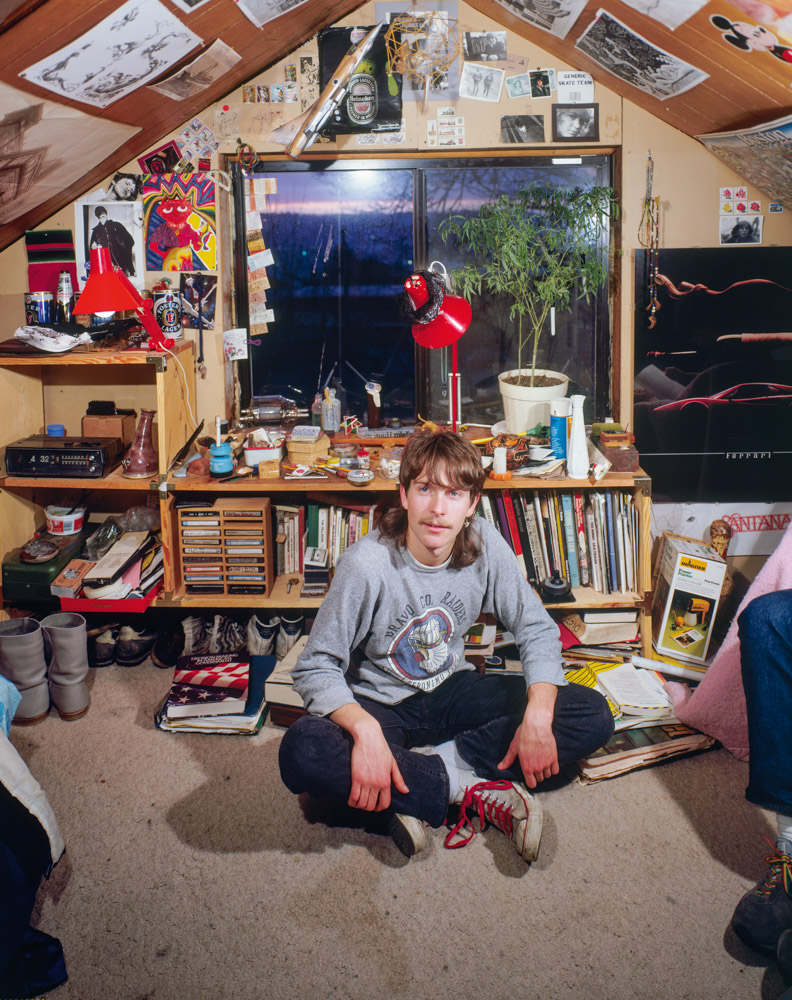 Person sitting cross-legged in cluttered attic room with sloped ceiling, shelves of books, red desk lamp, and papers pinned to walls.