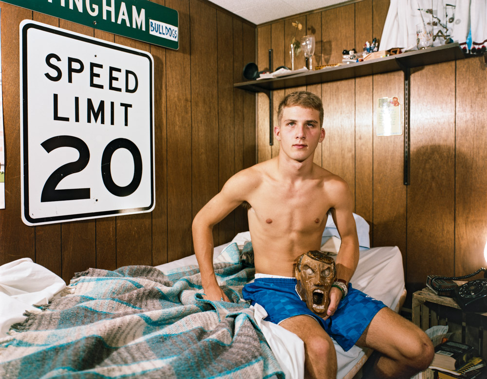 Shirtless young man in blue shorts sits on bed in wood-panelled room with speed limit 20 sign on wall