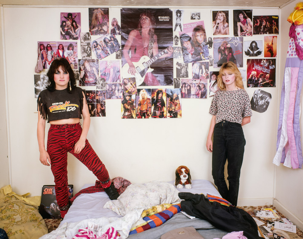 Two women posing in bedroom with posters covering wall behind them, clothes scattered on floor, small dog sitting between them.
