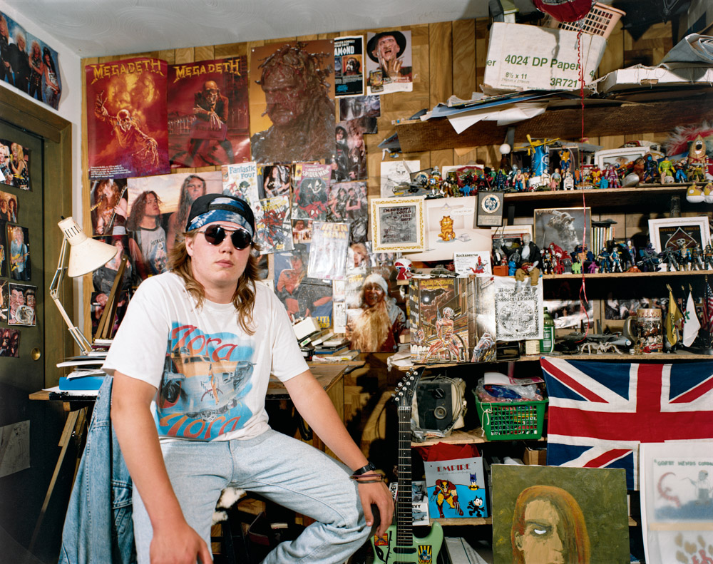 Person in sunglasses and white t-shirt sitting in cluttered room filled with posters, records, books and Union Jack flag.