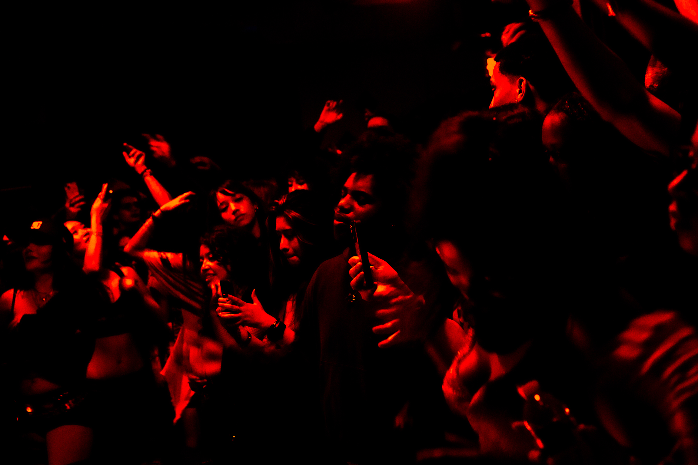 Crowd of people illuminated in dramatic red lighting against black background, creating high contrast silhouettes and shadows.