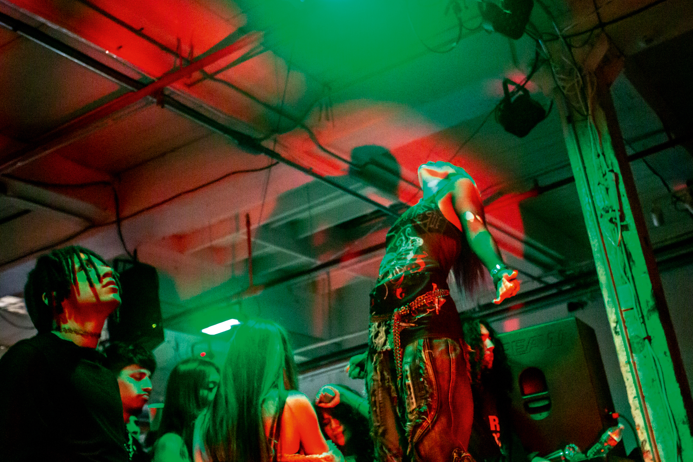 Performers in colourful traditional costumes under bright green and red stage lighting in an indoor venue with exposed ceiling beams.