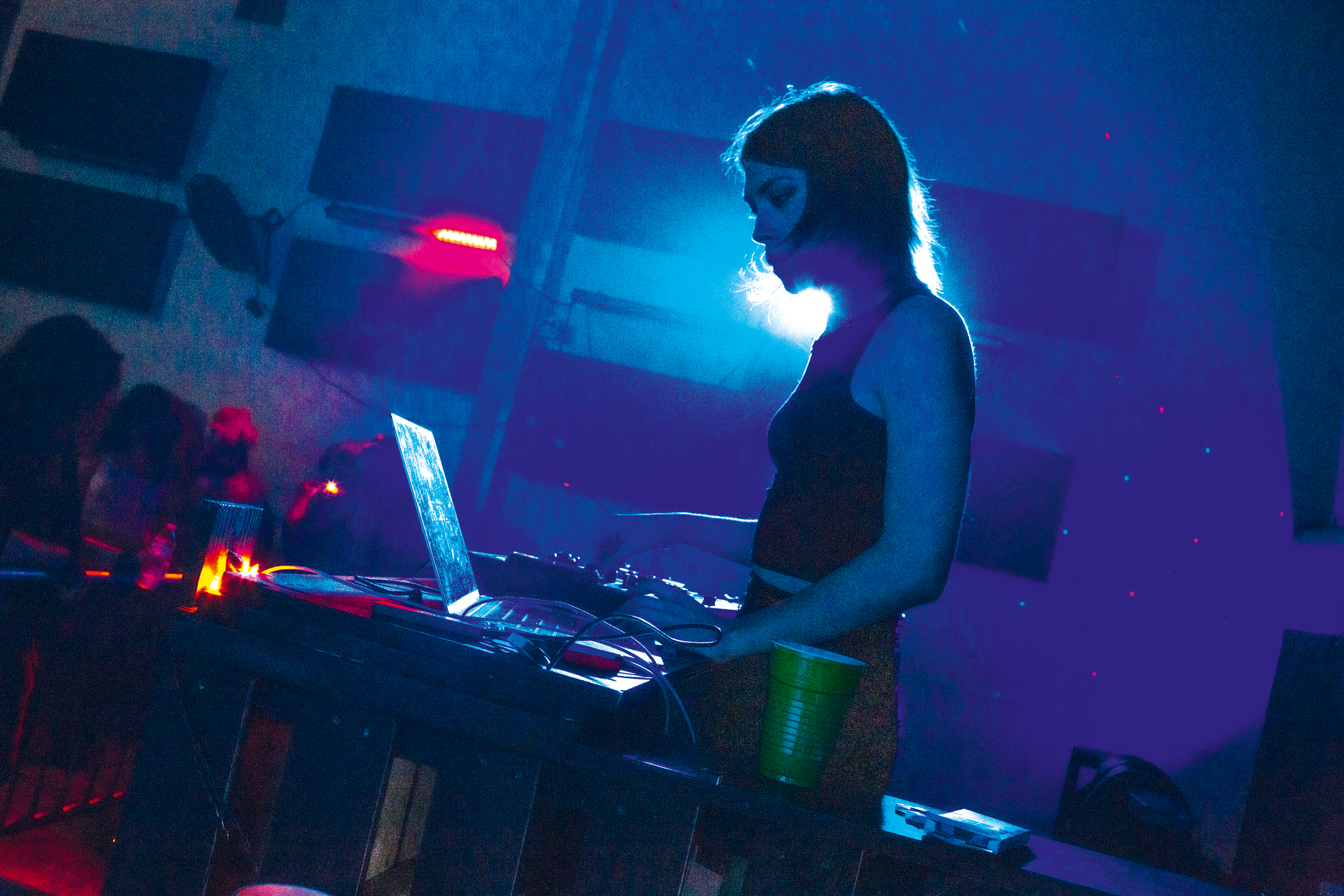Woman using laptop at desk in dimly lit room with blue and red neon lighting creating atmospheric glow.