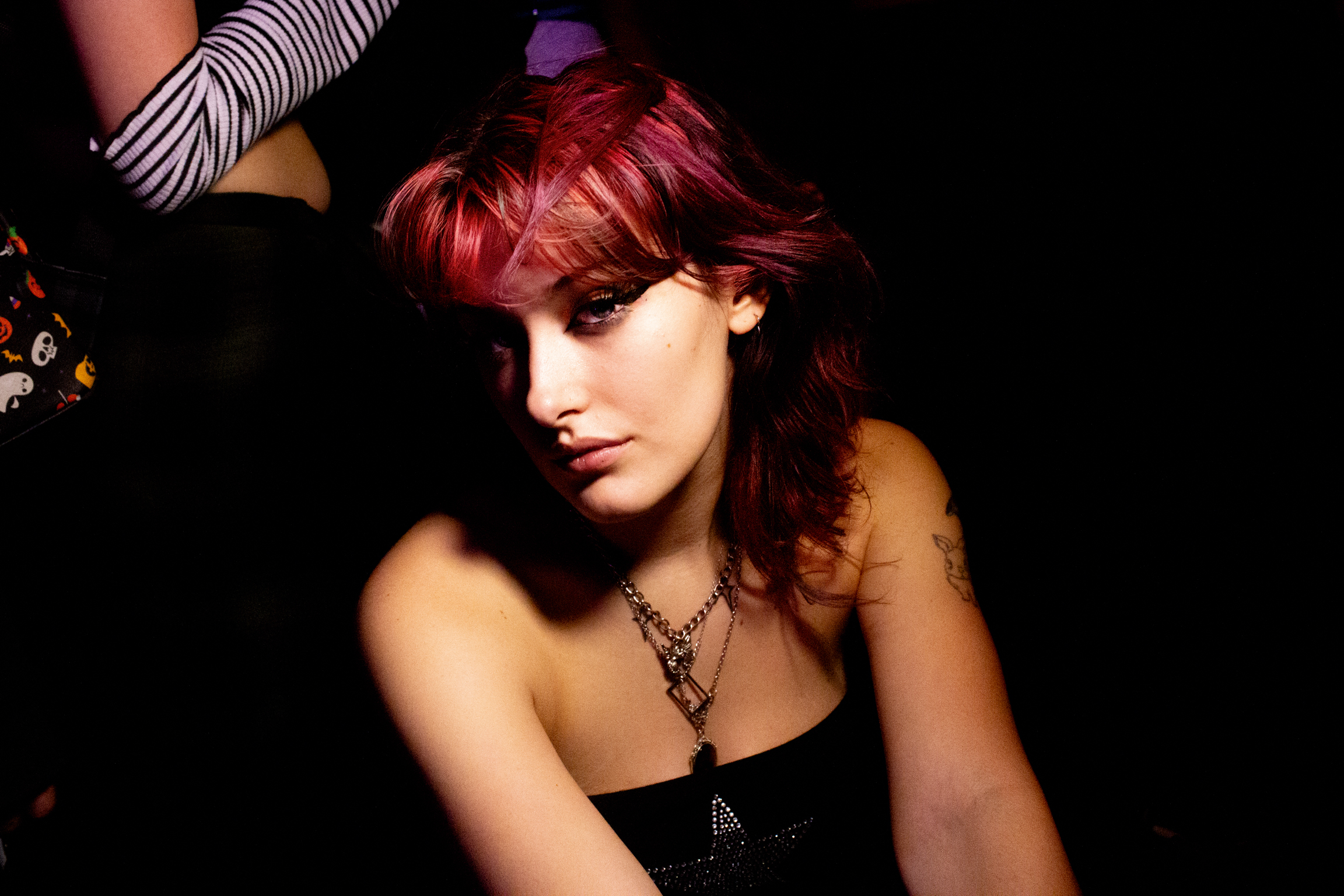 Woman with red hair and fringe wearing black strapless top, arms raised, against dark background with striped fabric visible.