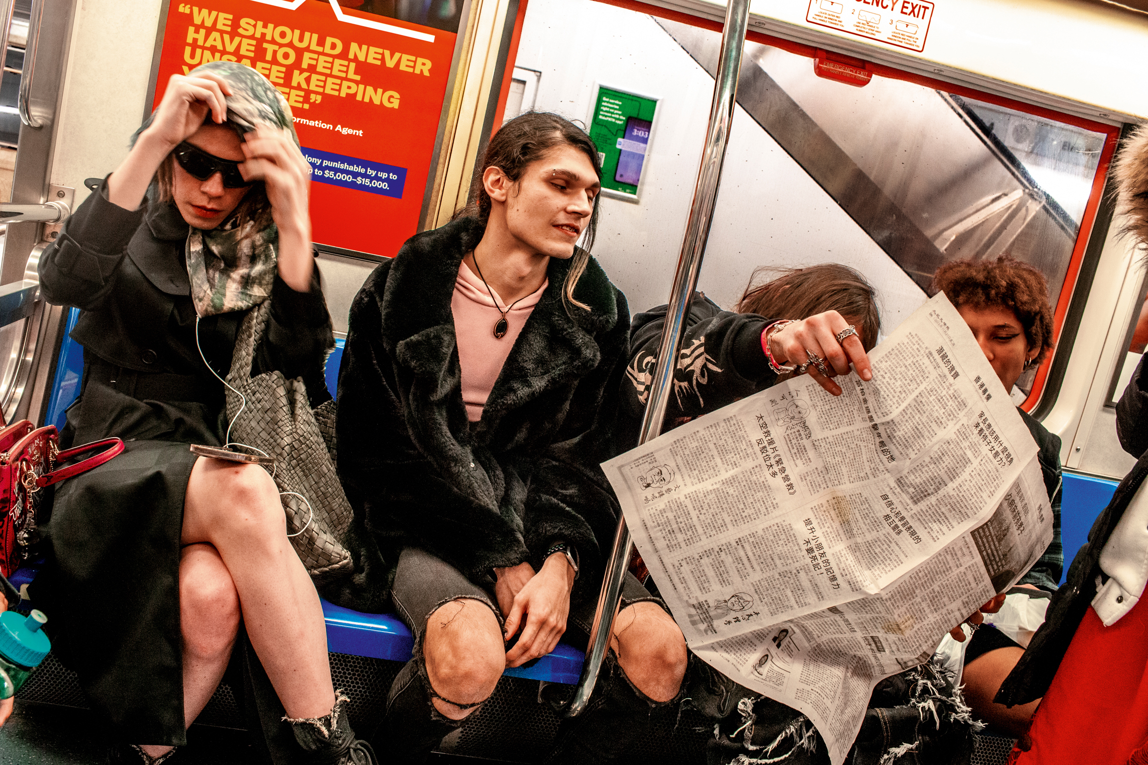 Three people sitting on blue tube train seats. Woman in sunglasses and scarf, woman in black coat, man reading newspaper. Red safety poster visible.